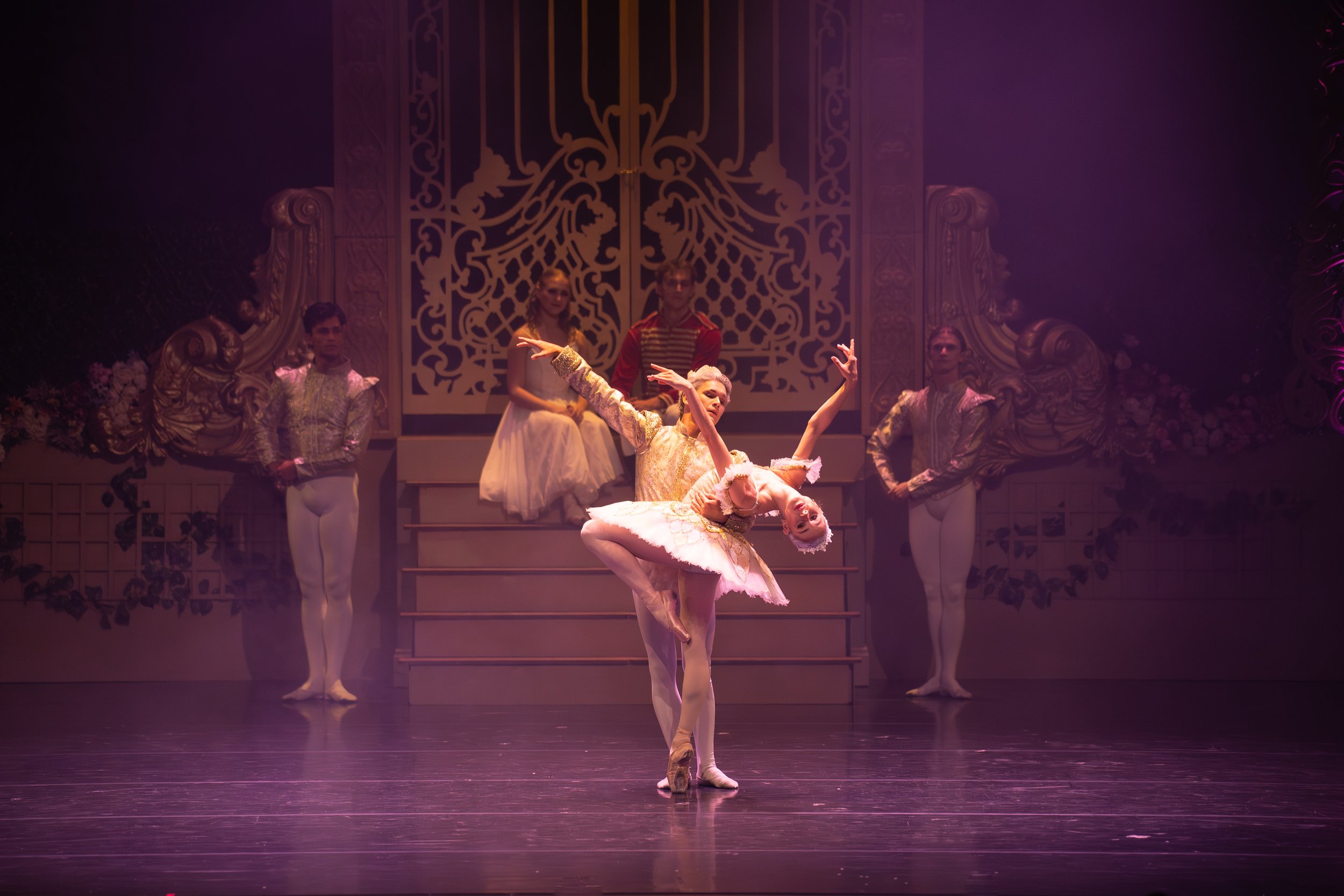Ballet dancers perform on stage with ornate backdrop, with one dancer in front en pointe and another leaning back with arms raised.