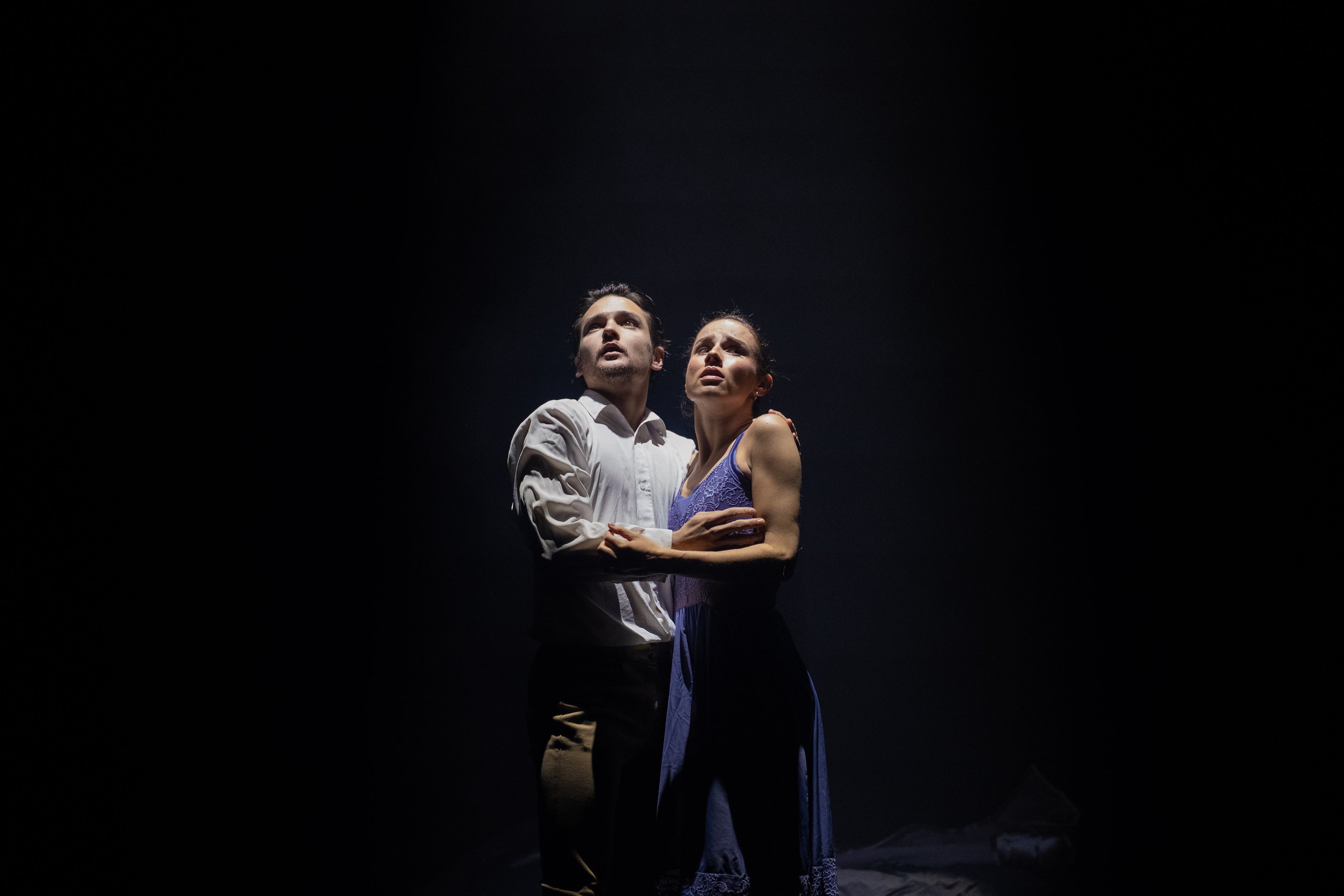 A man and woman with serious expressions embrace in a dark setting, illuminated from below.