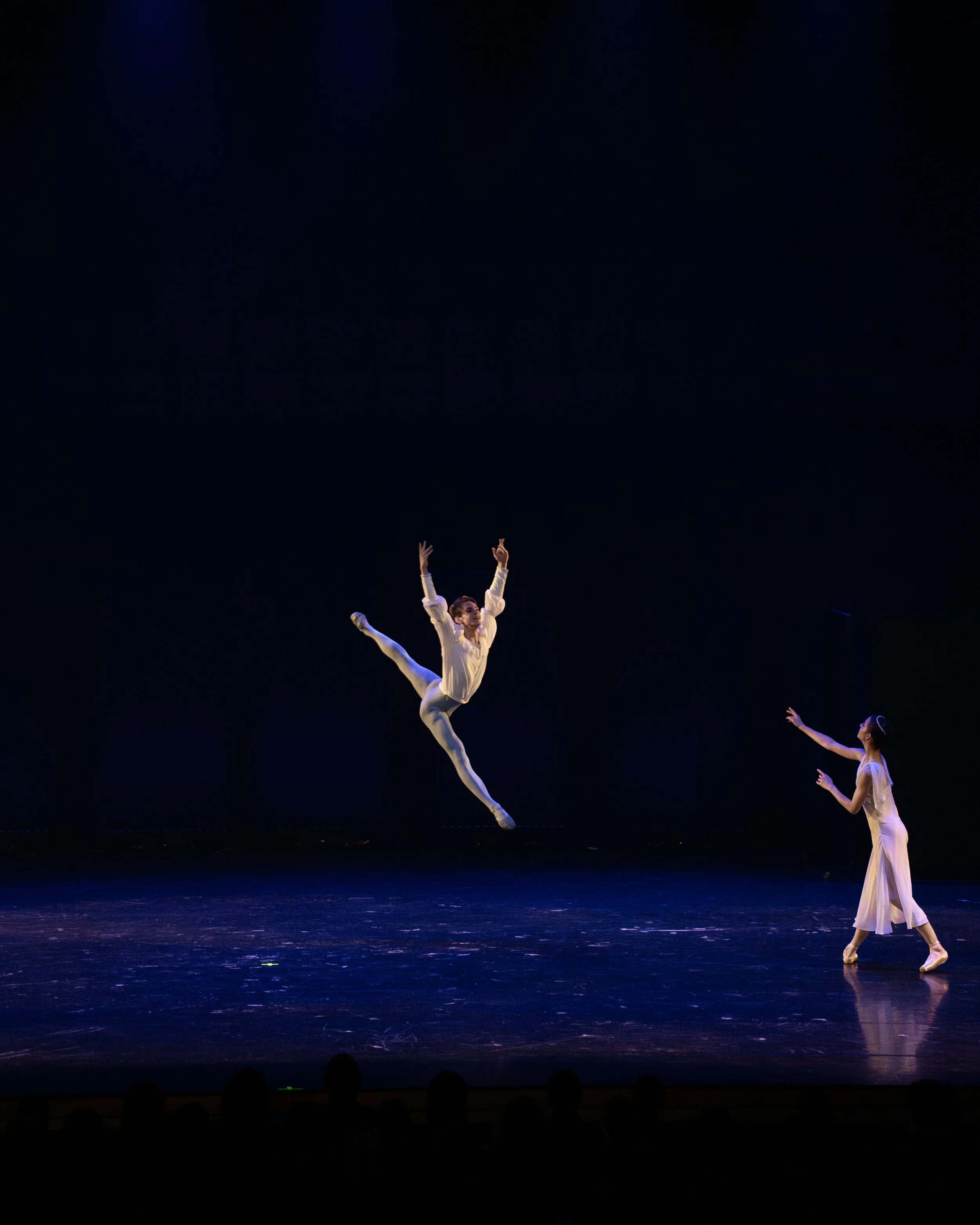 Ballet dancers perform on stage, one dancer mid-air in an arabesque, while another stands on the stage with outstretched arms, all dressed in light-colored costumes, under dim stage lighting.