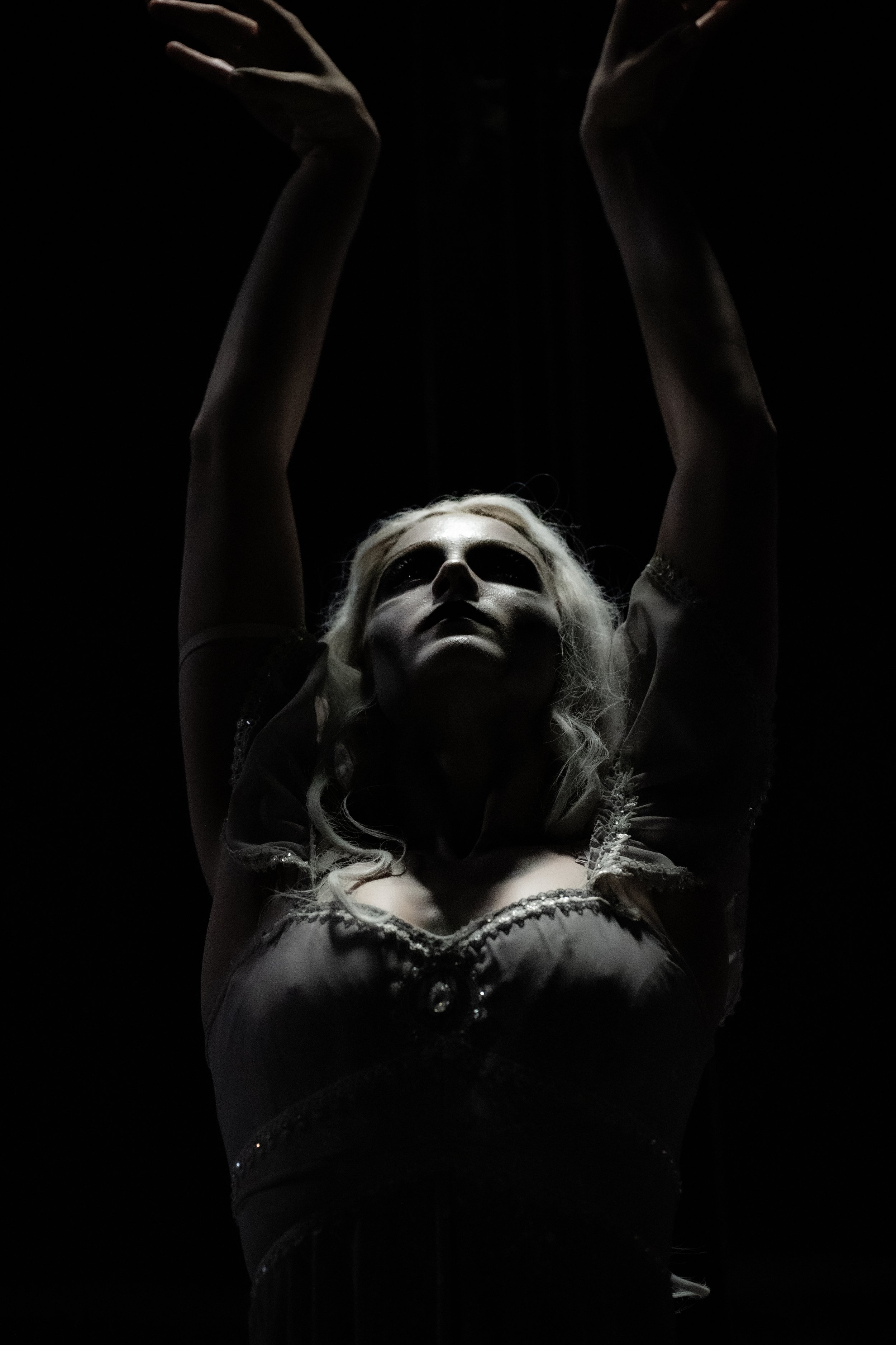 A person with long, wavy, platinum blonde hair is raising their arms above their head, with hands poised as if performing a dance or pose, in a dark setting with subtle lighting highlighting their face and upper body.