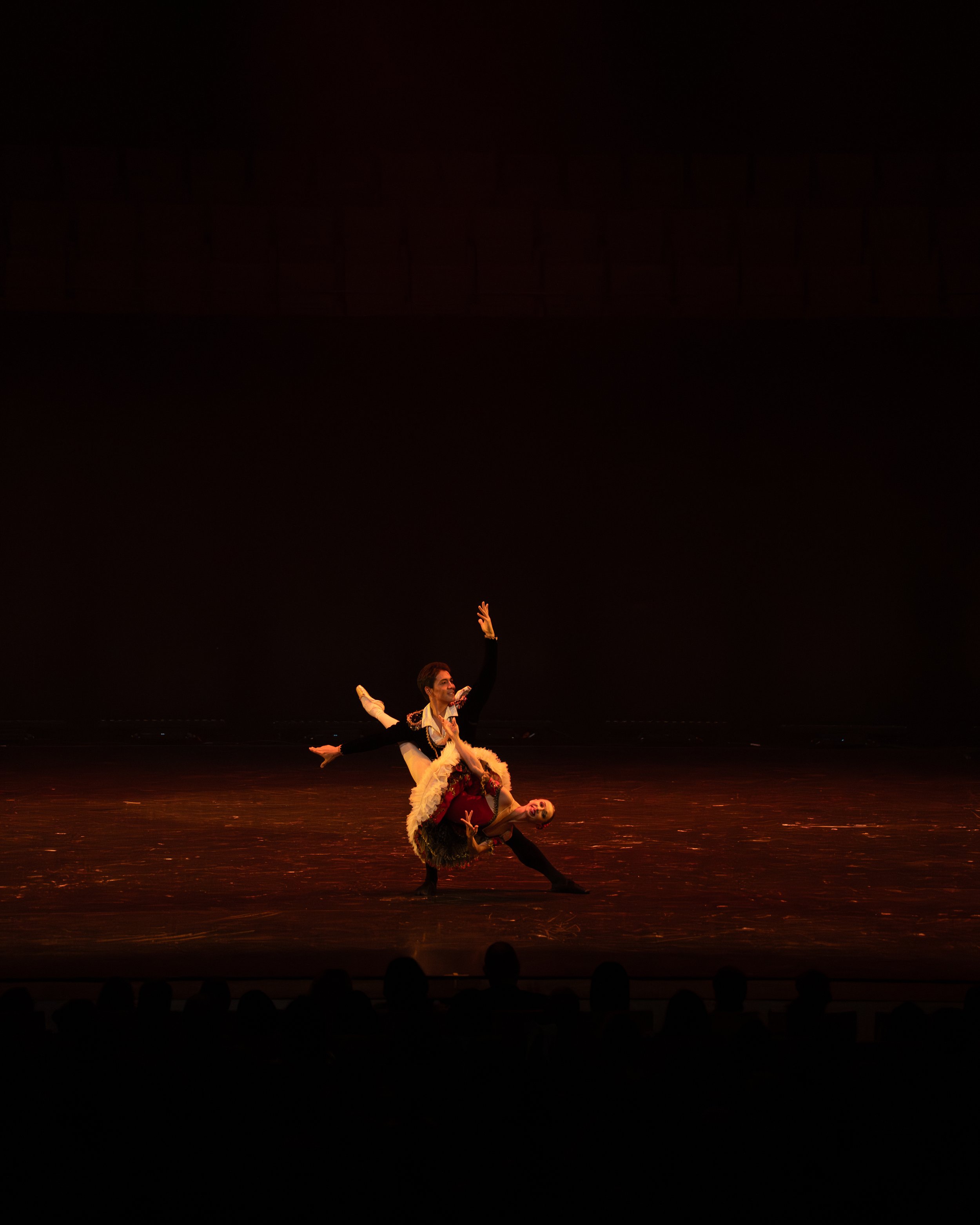 Two ballet dancers perform on stage with a dark background, one standing with arms raised and the other lying on the floor, both dressed in elaborate costumes.