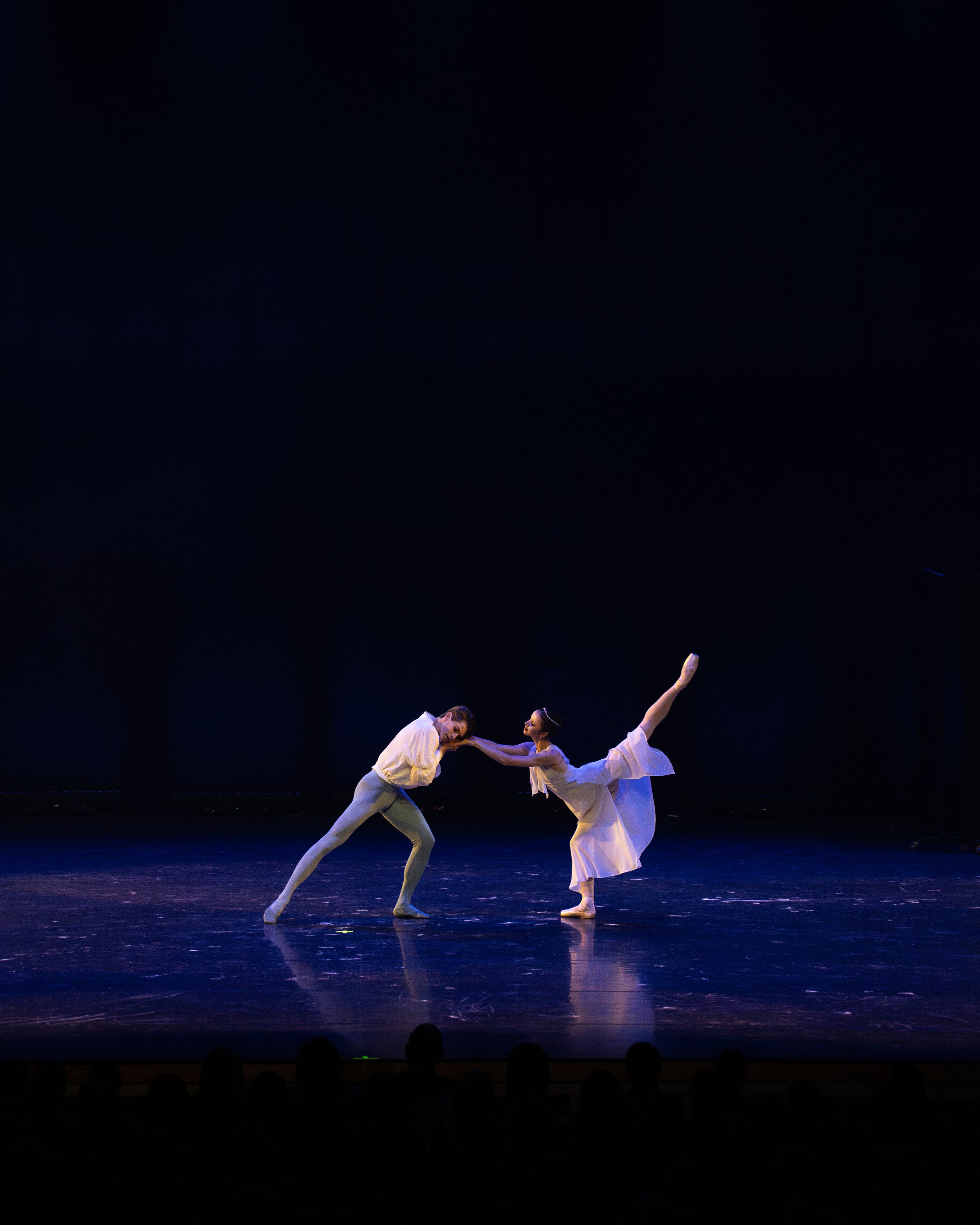 Two ballet dancers performing on stage, with one dancer supporting the other in a high arabesque pose, both wearing white costumes against a dark background.