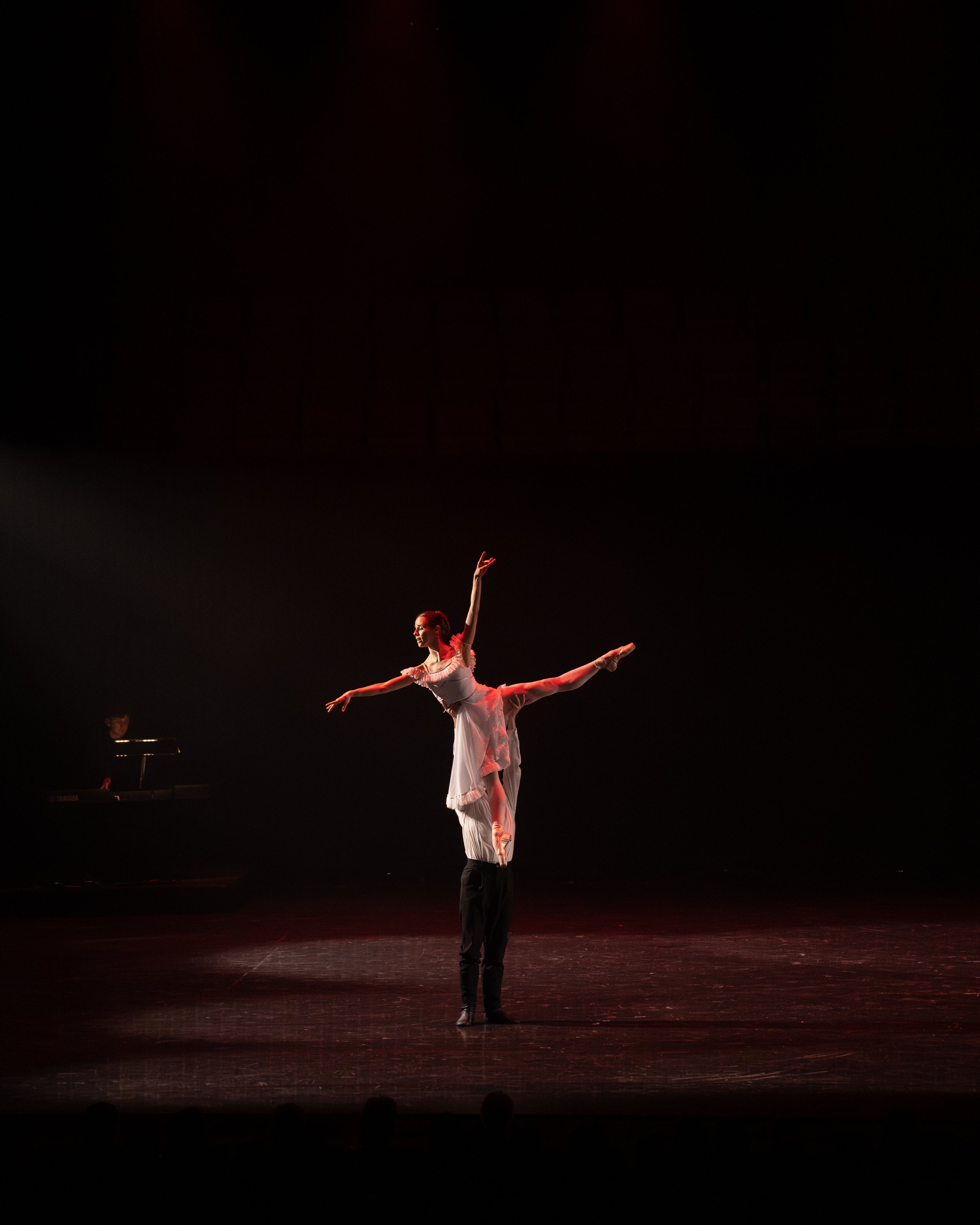 Ballet dancer performing on stage with a male partner supporting her in a pose, stage is dark with minimal lighting.