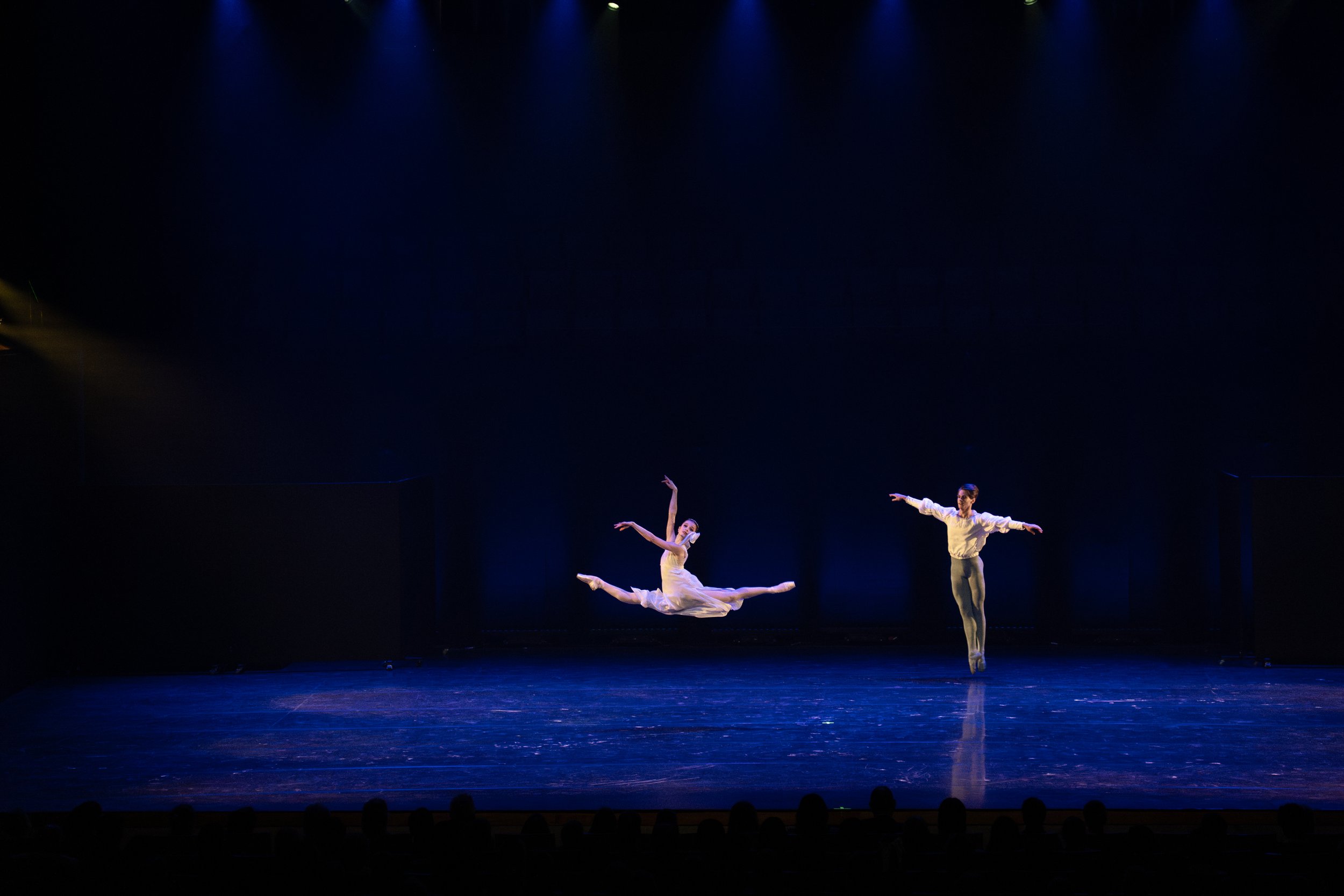Three ballet dancers performing on a dark stage with blue spotlighting. One dancer is in midair during a leap, two others stand on the stage with arms extended.