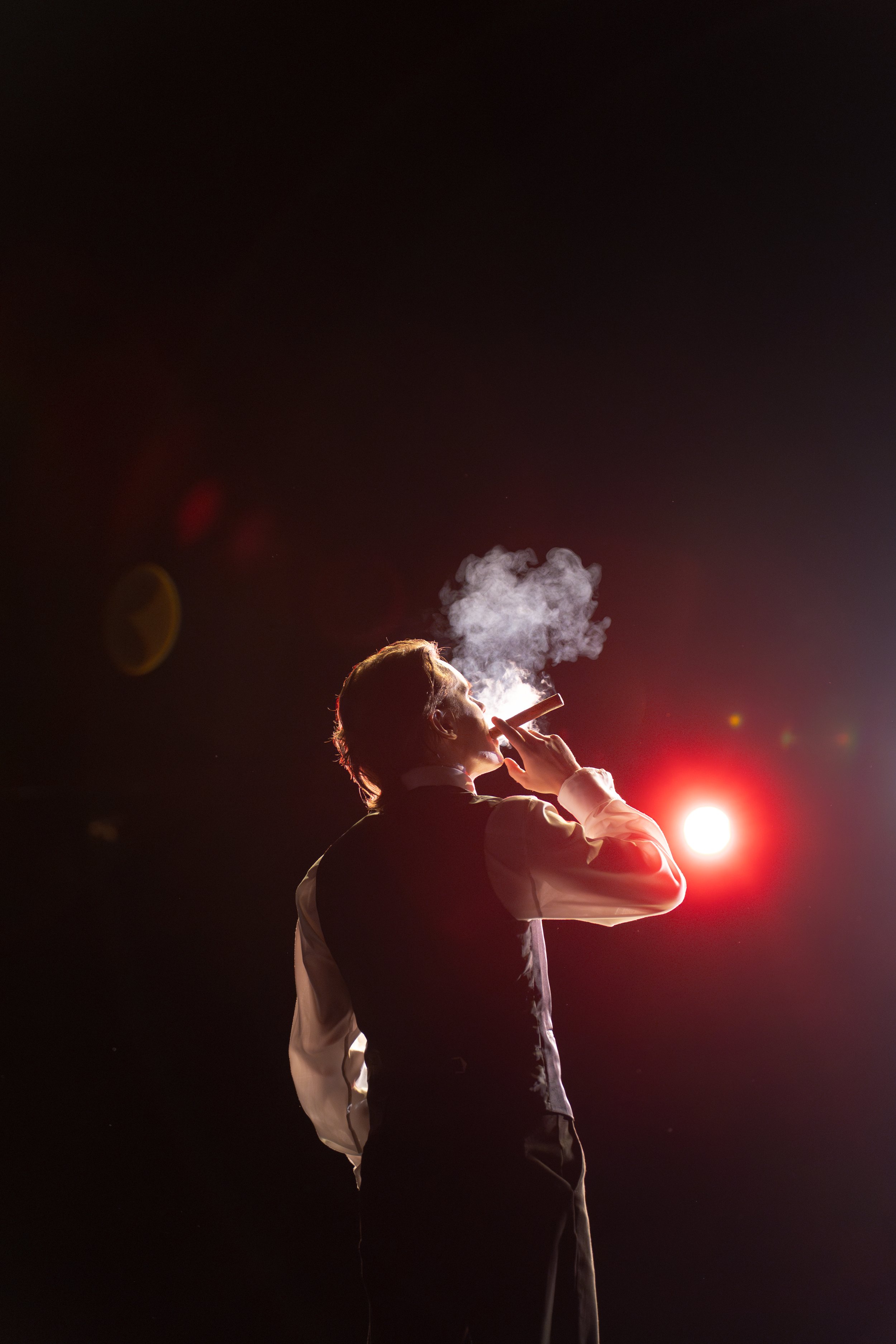 A person smoking a cigar outdoors at night, with smoke rising from their mouth, illuminated by a bright light behind them.