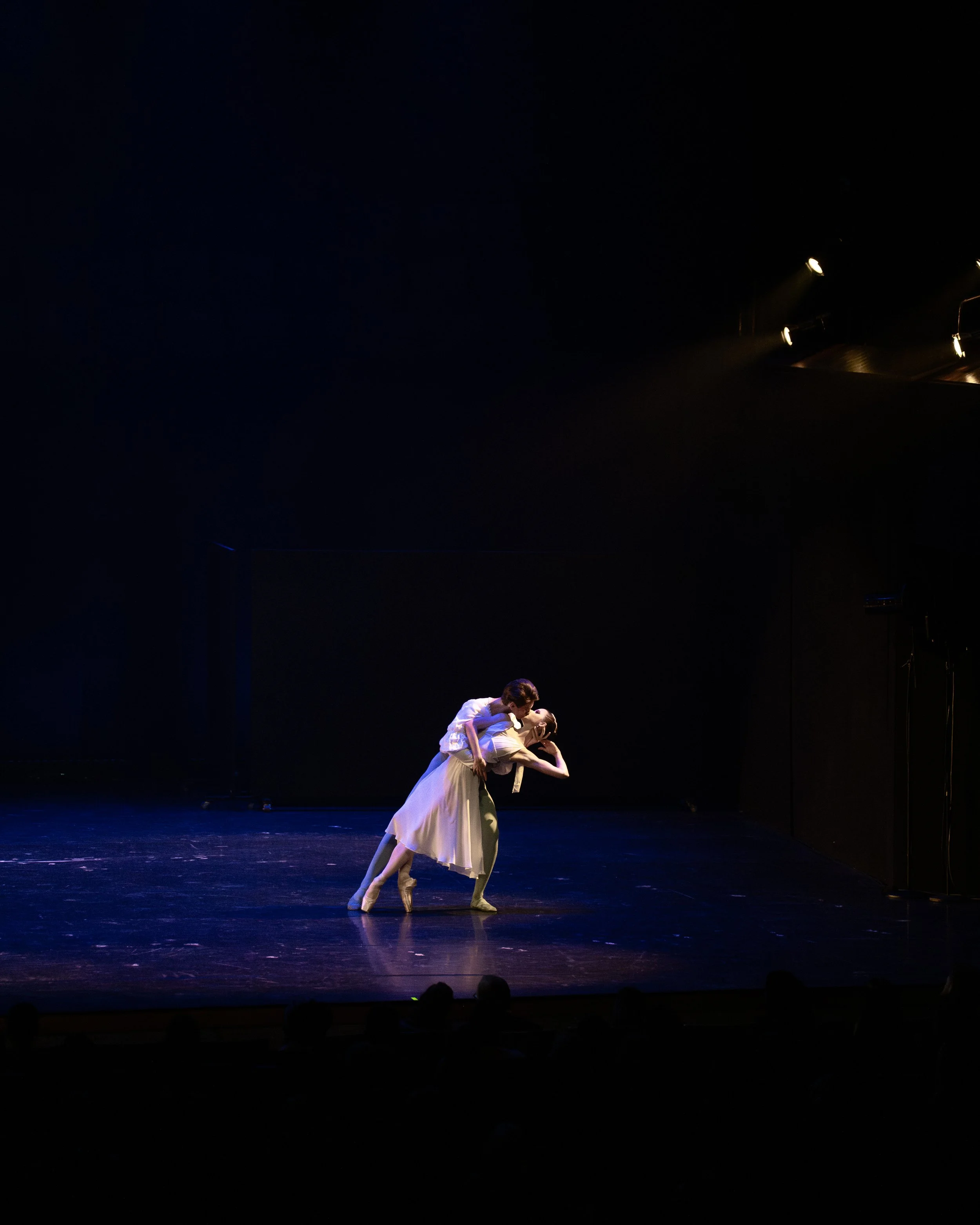 Two ballet dancers performing on stage under dim lighting, with one dancer supporting the other in a lift, both dressed in light-colored costumes.