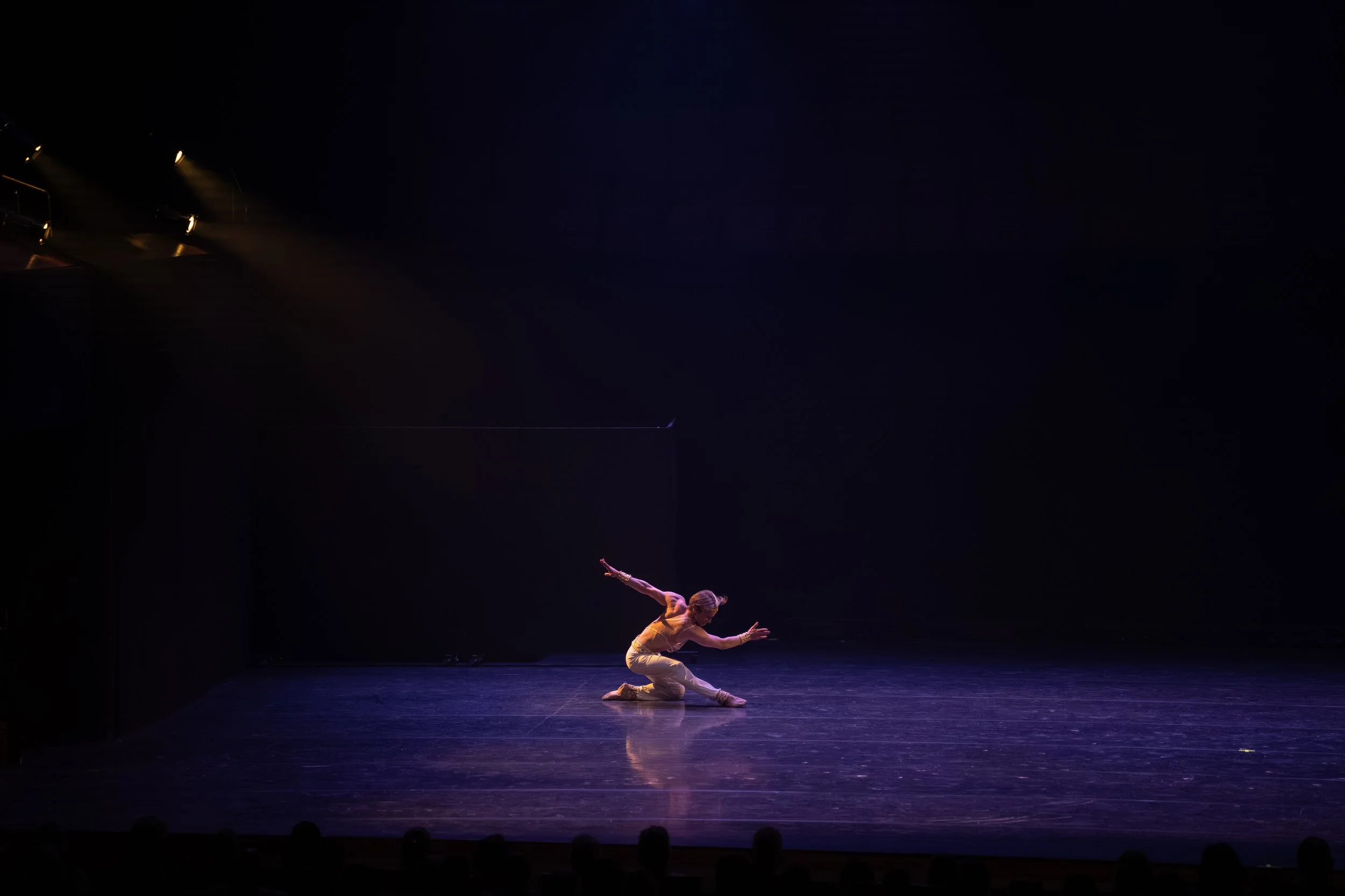 A performer in a yellow costume is kneeling on stage with arms extended outward, performing a dance or acrobatic move under stage lighting.