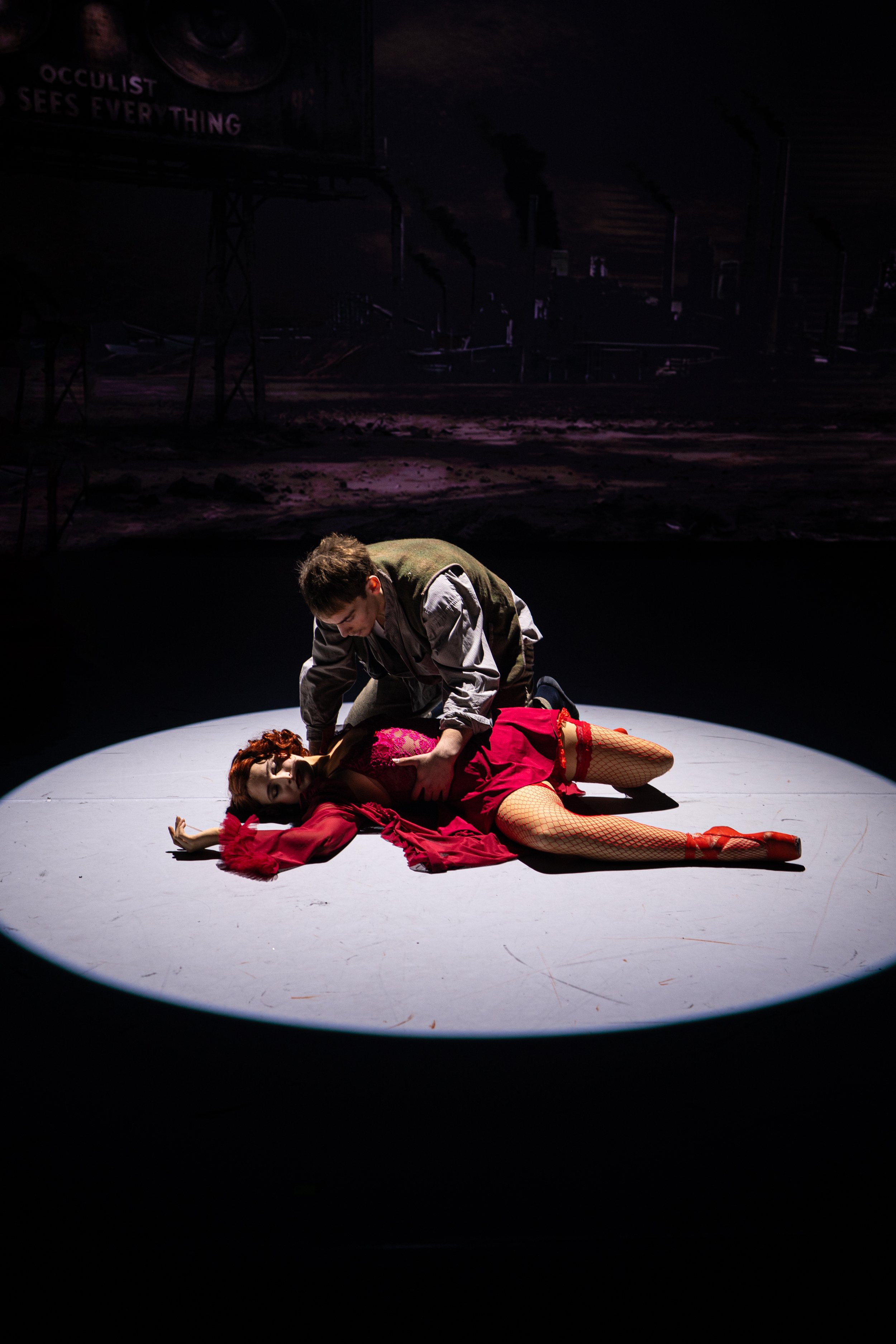 A theatrical scene with a performer lying on the stage in a red dress and fishnet stockings, and another performer kneeling next to her. The scene is lit within a circular spotlight, with a dark background and a large screen or backdrop behind them.