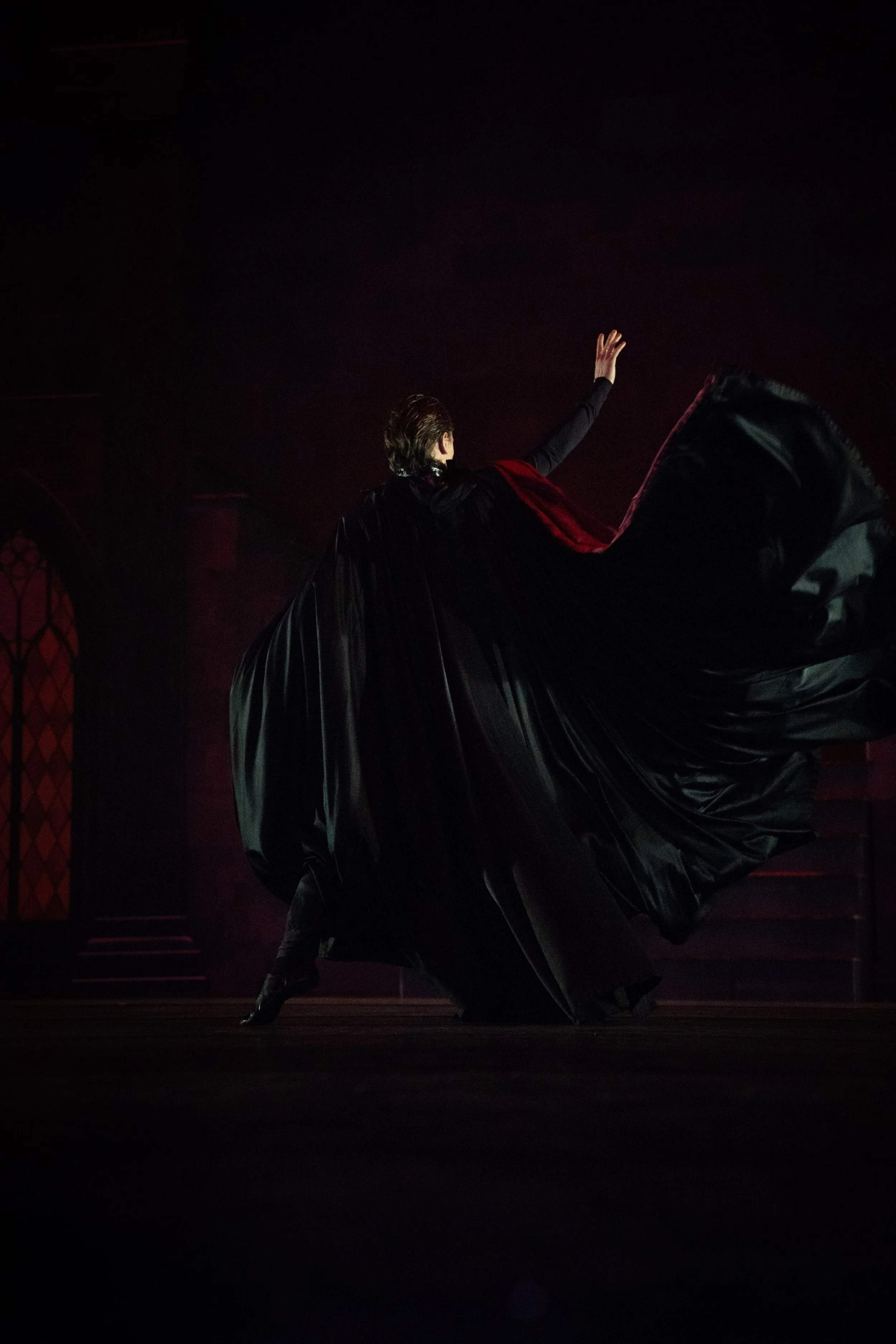 A male flamenco dancer performs on a dimly lit stage, wearing a black outfit with a large, flowing black skirt and a red sash or cape, with his arm raised and one foot extended.
