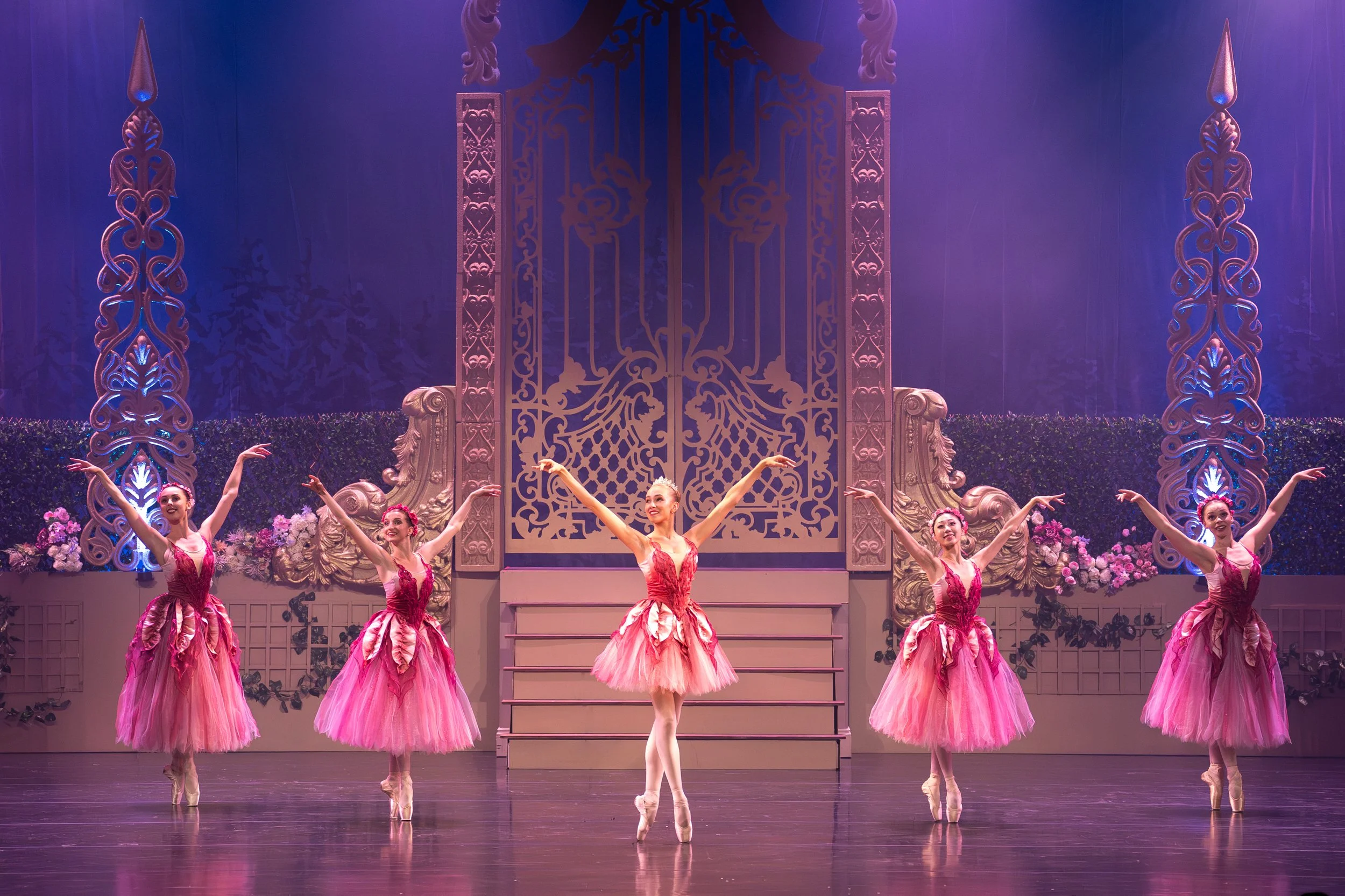 Ballet dancers in pink dresses performing on stage with a decorated backdrop and floral arrangements.