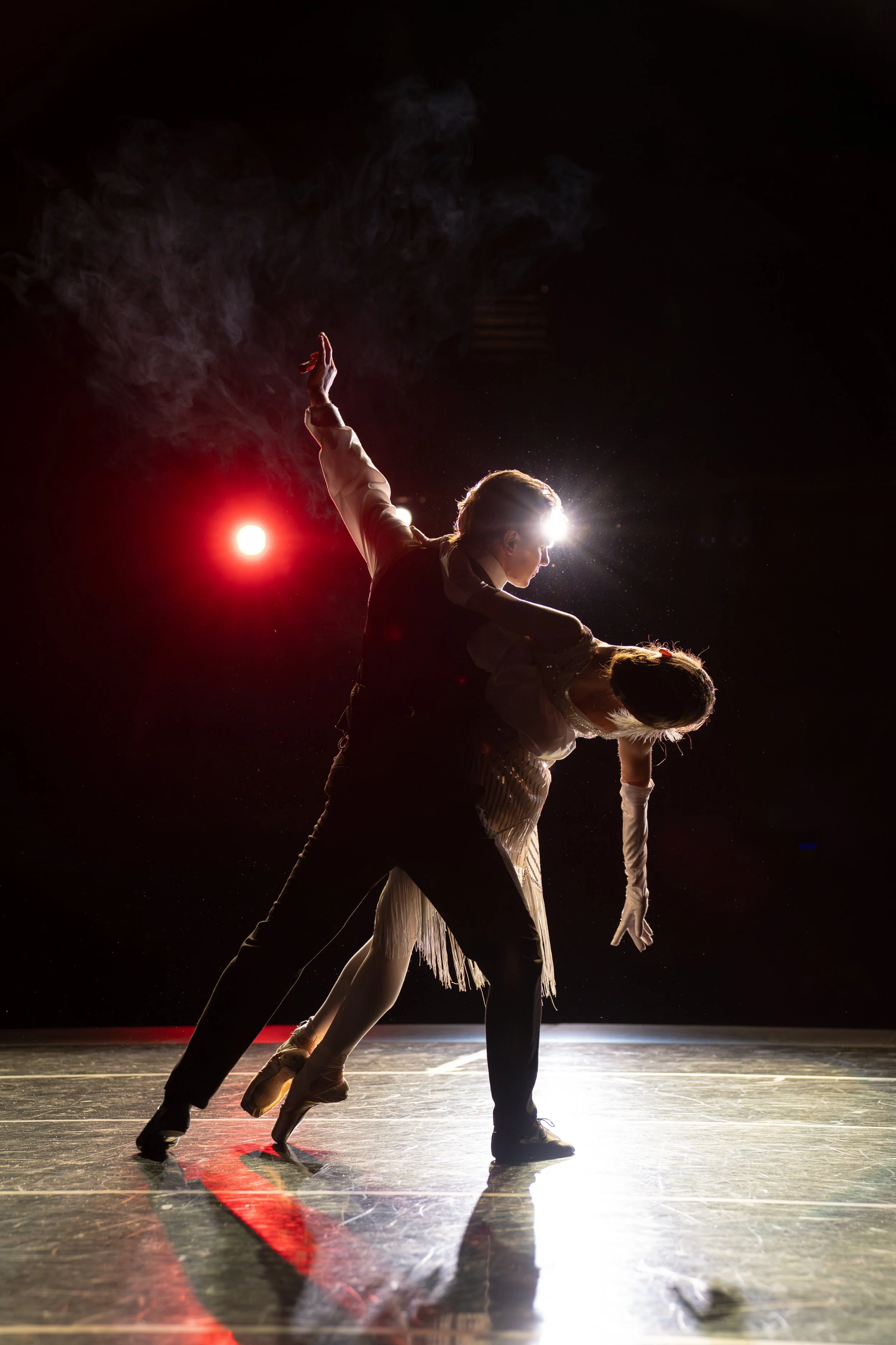 Two ballet dancers performing on stage with dramatic lighting, one dancer supporting the other in a pose, with smoke and a bright light behind them.