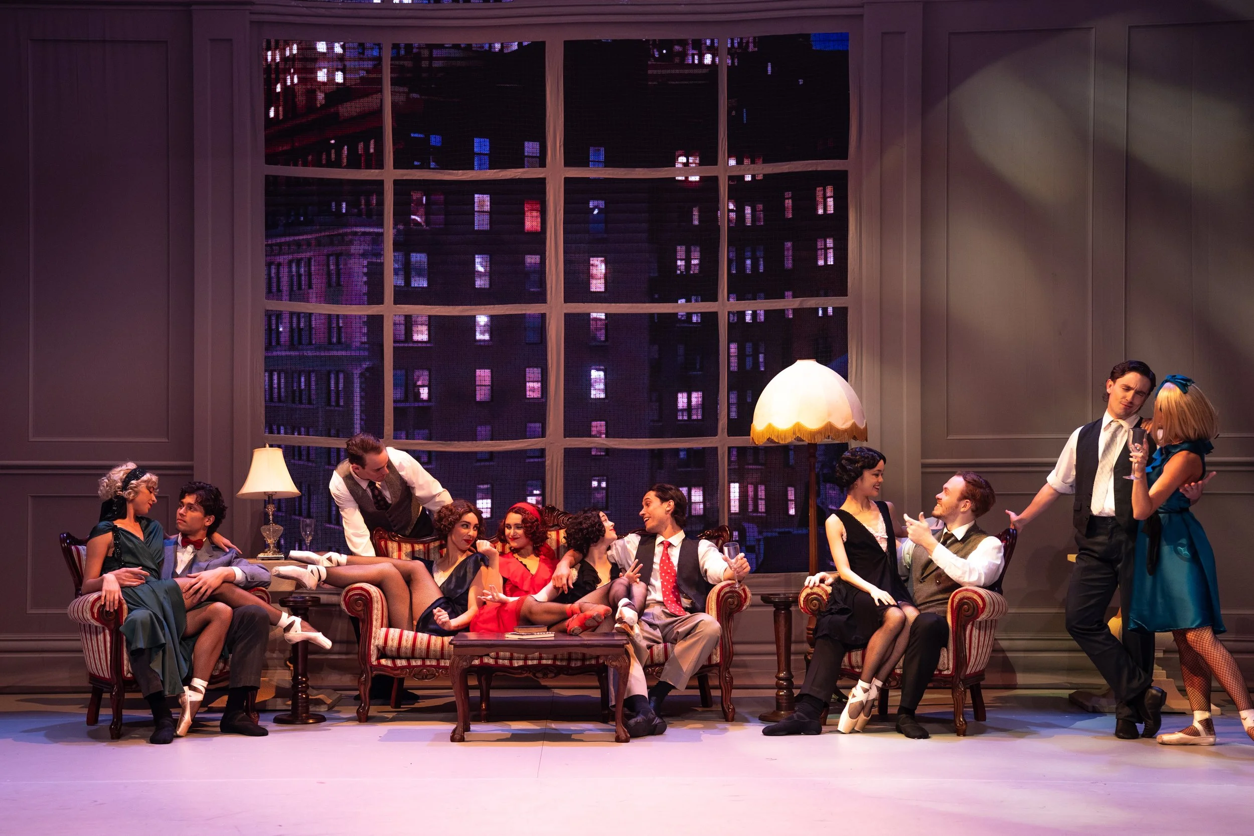 Stage scene from a musical or play set in an elegant living room with large window showing city buildings at night. Several well-dressed men and women are sitting and standing, engaging in conversation and laughter, with some holding drinks. The ligh