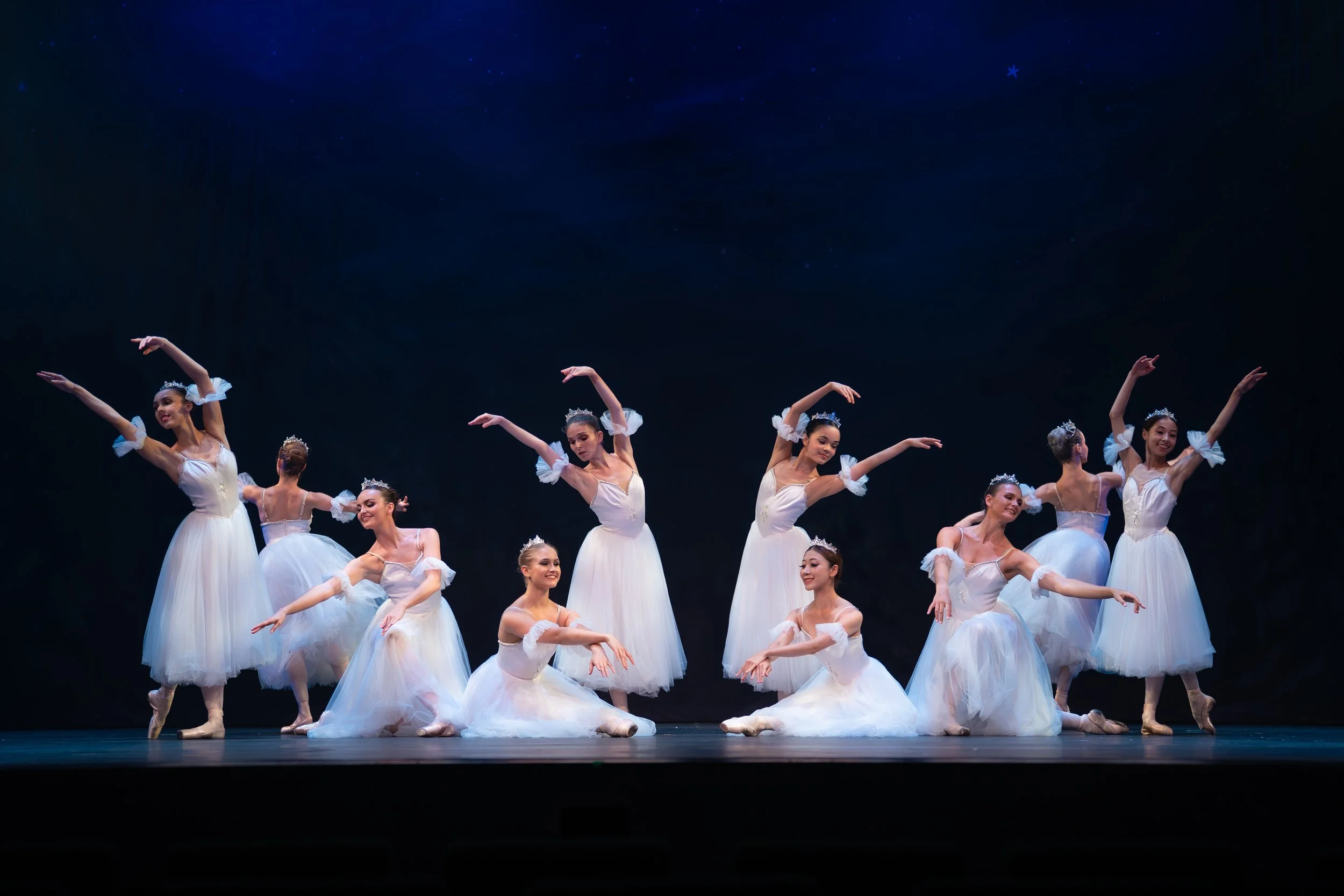 Ballet dancers in white costumes performing on stage, with dark background and stars projected above.