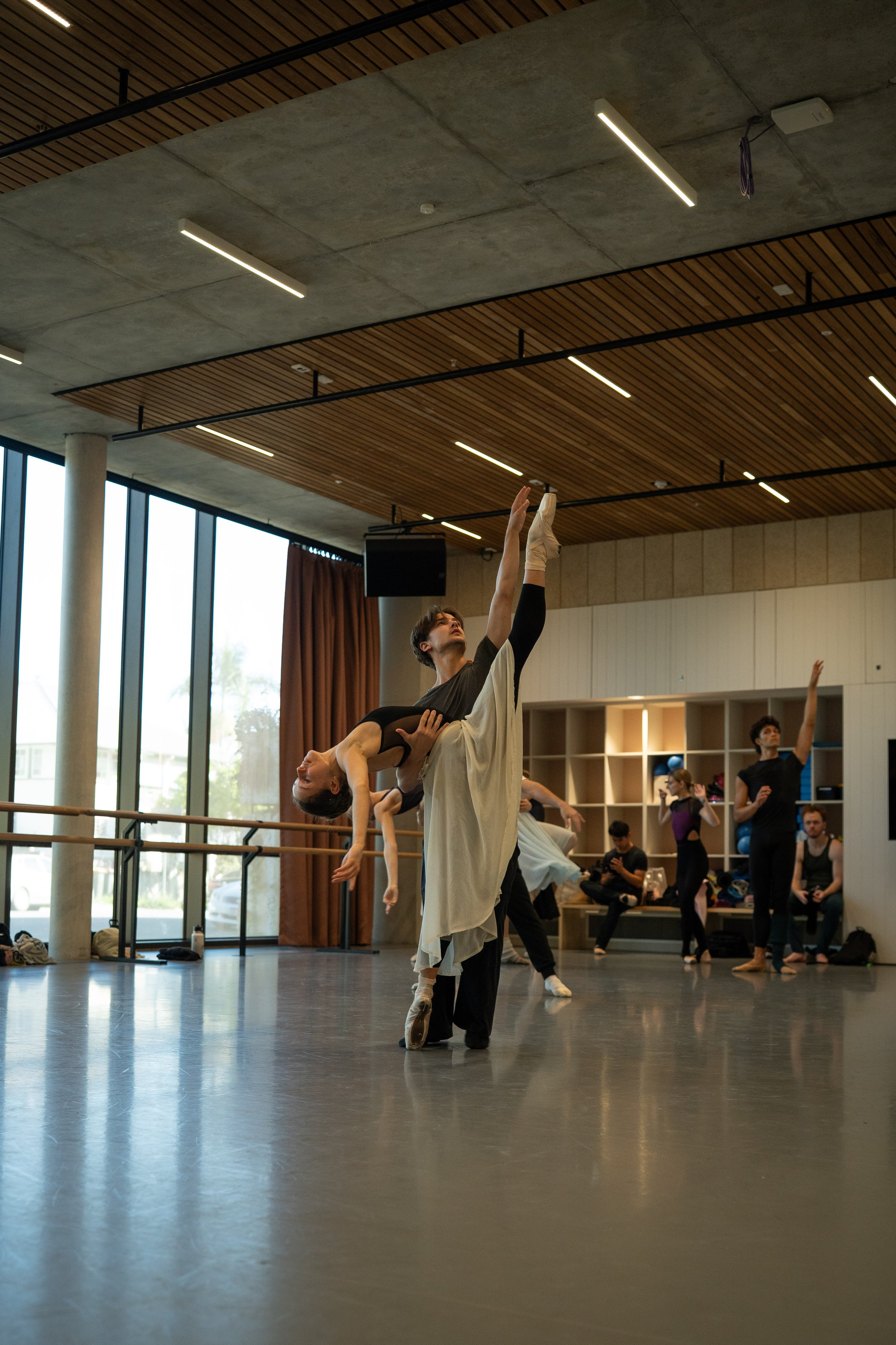 Ballet dancers practicing in a dance studio with wooden and concrete ceiling, large windows, and people observing and taking photos in the background.