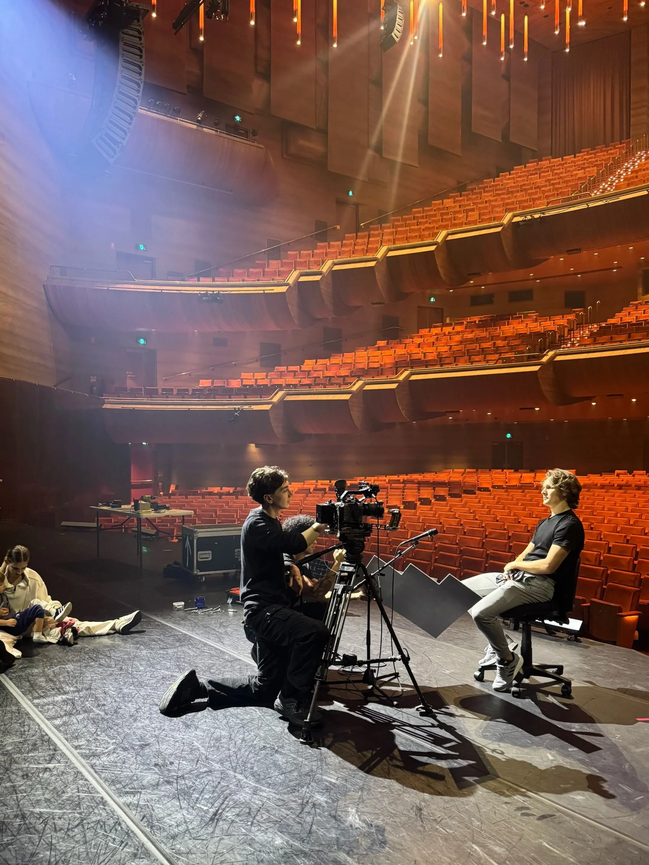 The image shows a theater stage with a camera operator filming a young man sitting on a chair, with rows of empty orange seats in the background. Several crew members are sitting on the stage, preparing for filming or rehearsal.