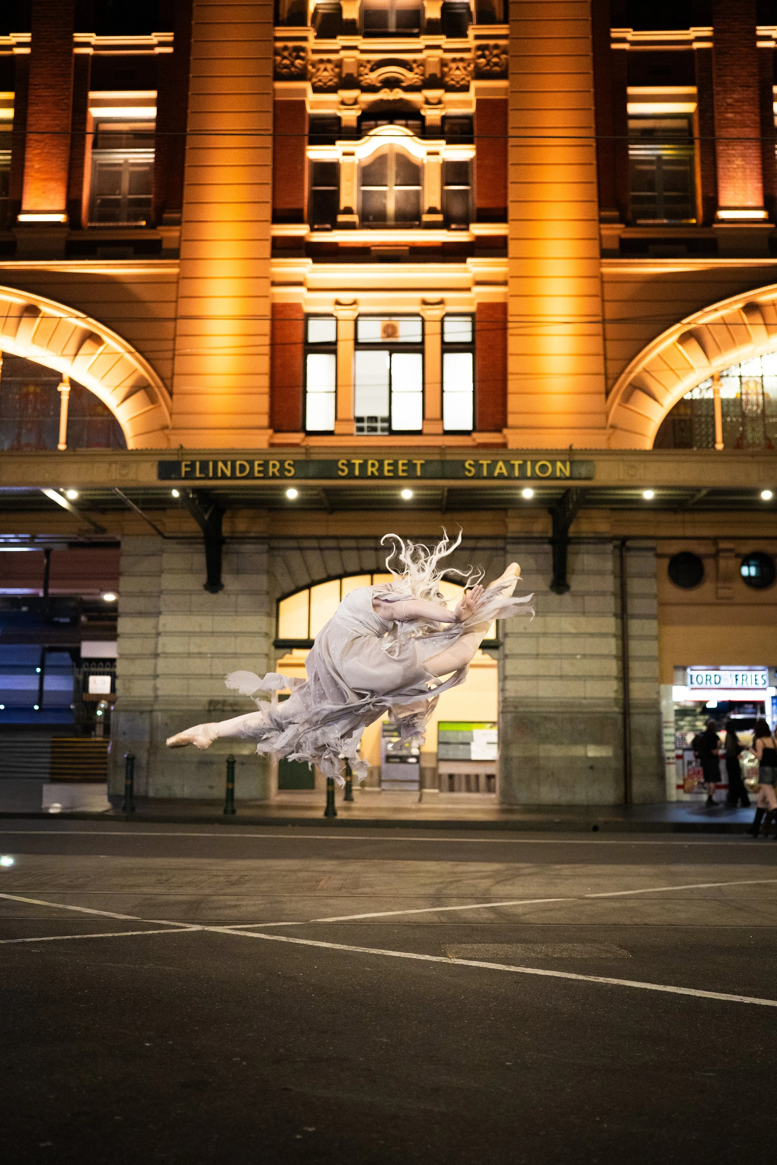 A dancer in a flowing white costume performing an aerial dance in front of a lit building at night, with the building's sign reading 'Flinders Street Station' in the background.