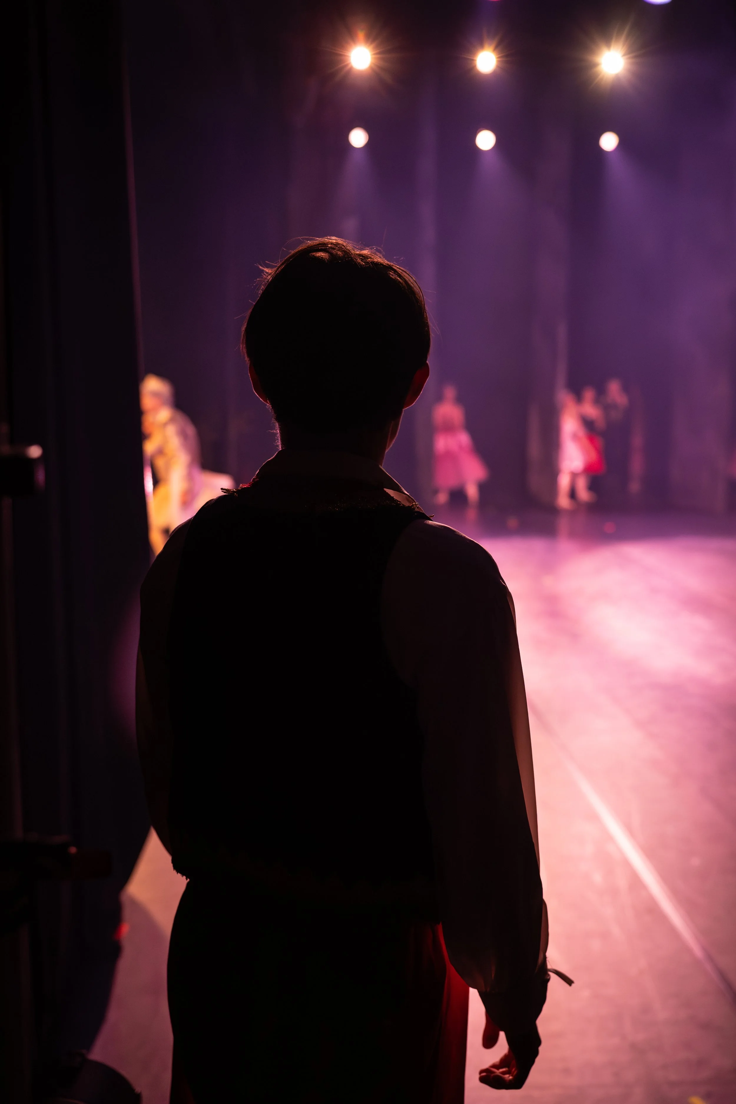 Backstage view of a performer watching others rehearse on stage with purple lighting.