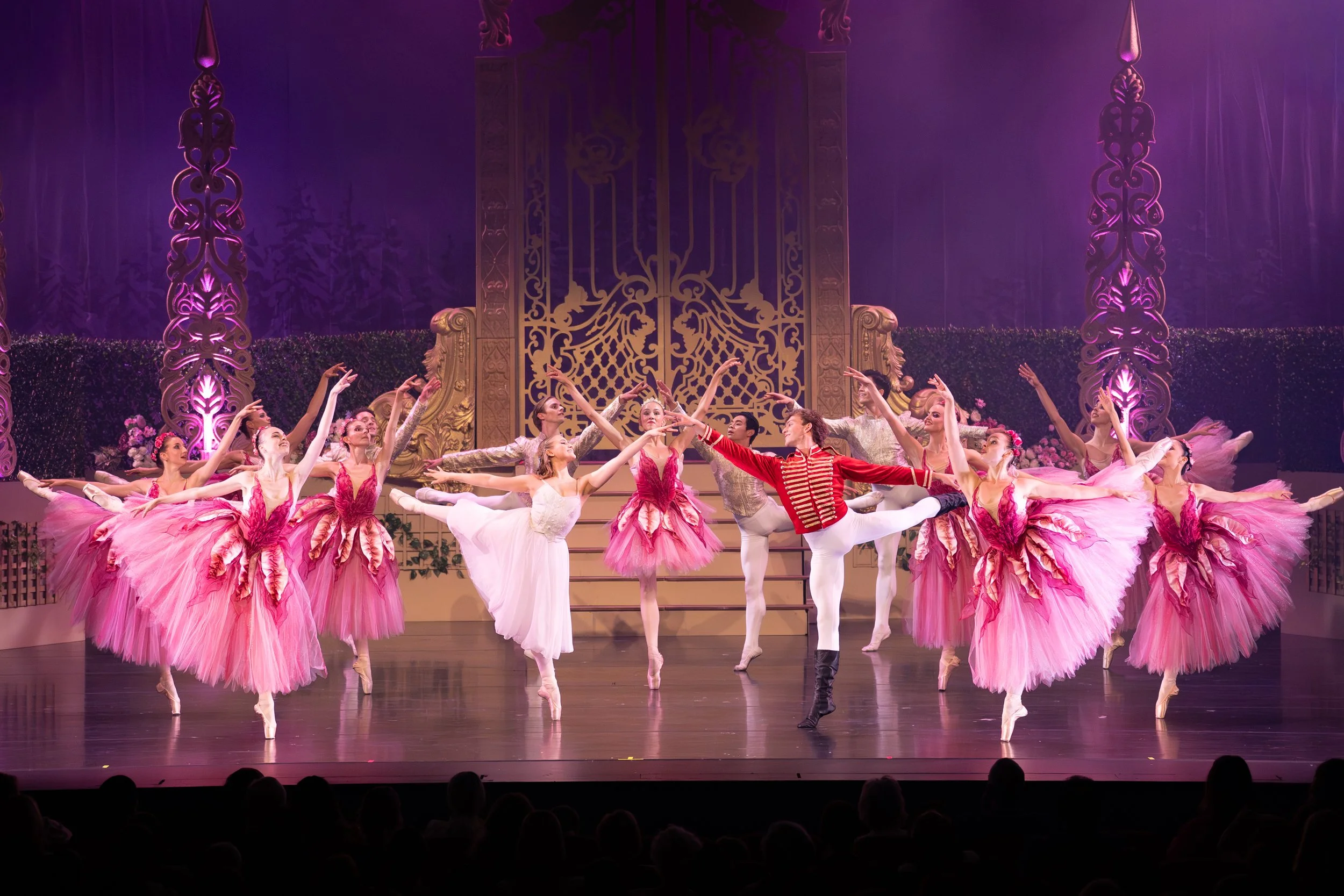 Ballet dancers performing on stage, many in pink tutus with floral accents, along with male dancers in tights and costumes, in an ornate theater with purple and gold decor.