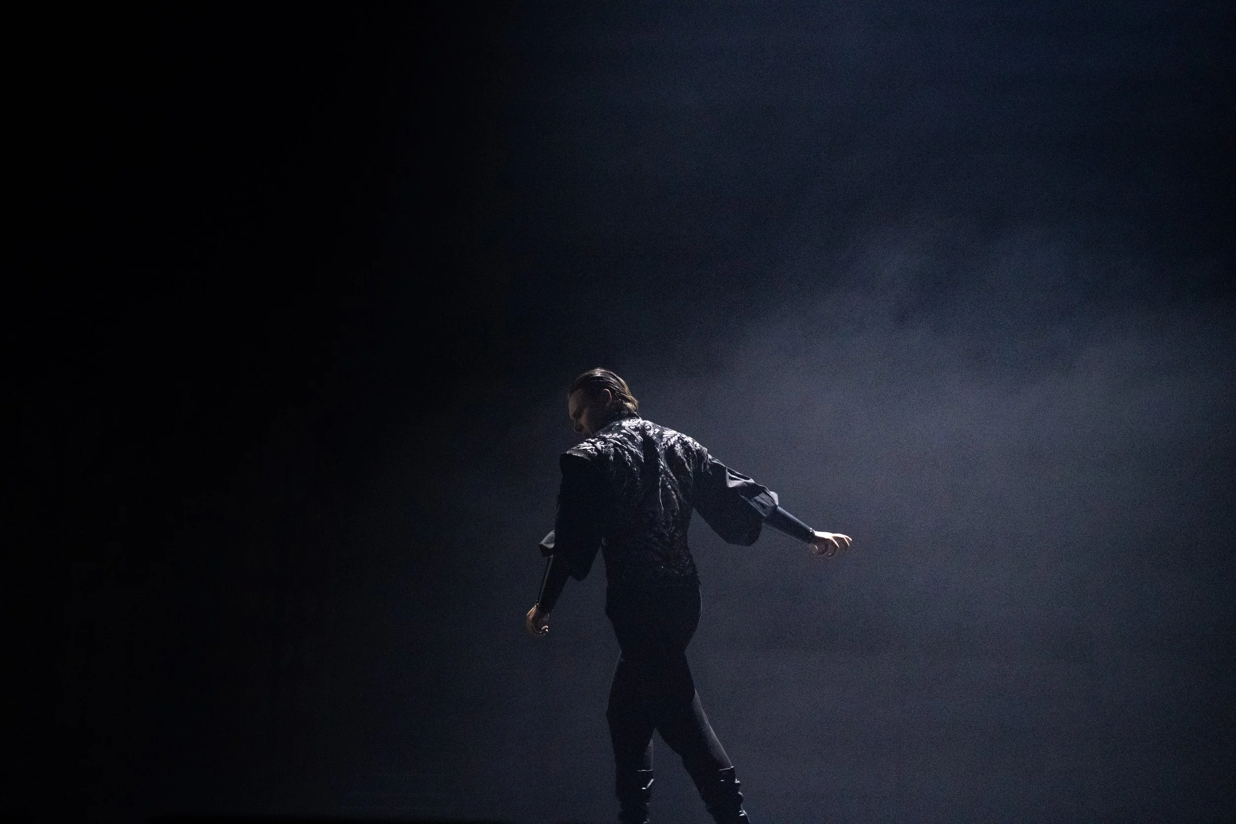 A person dressed in a patterned shirt and black pants is dancing on stage with one arm extended outward, dimly lit against a dark background.