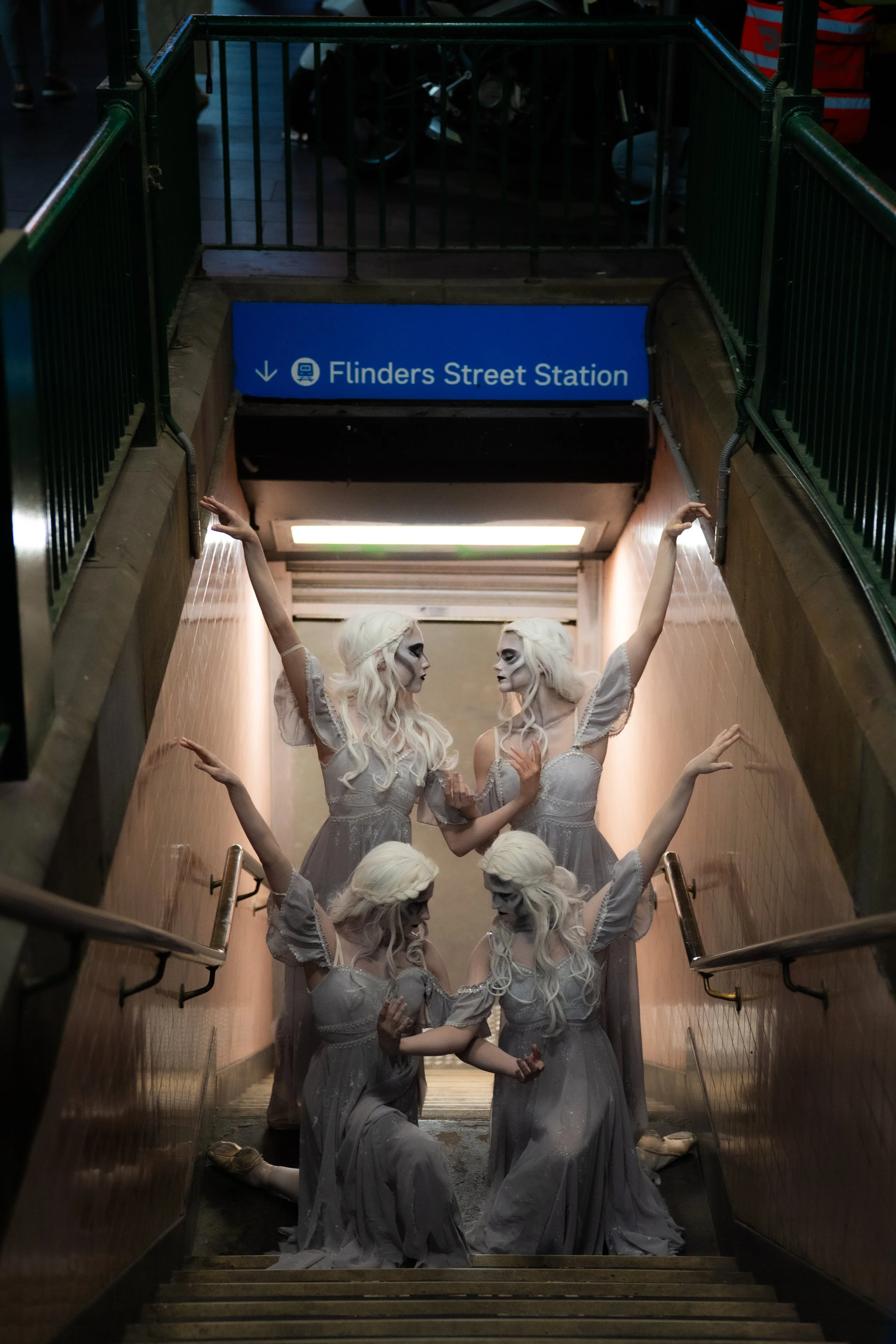 People dressed in grey historical costumes, with white wigs, posing on a staircase inside underground subway station, indicated by a blue sign with white writing that says "Flinders Street Station" and a downward arrow.