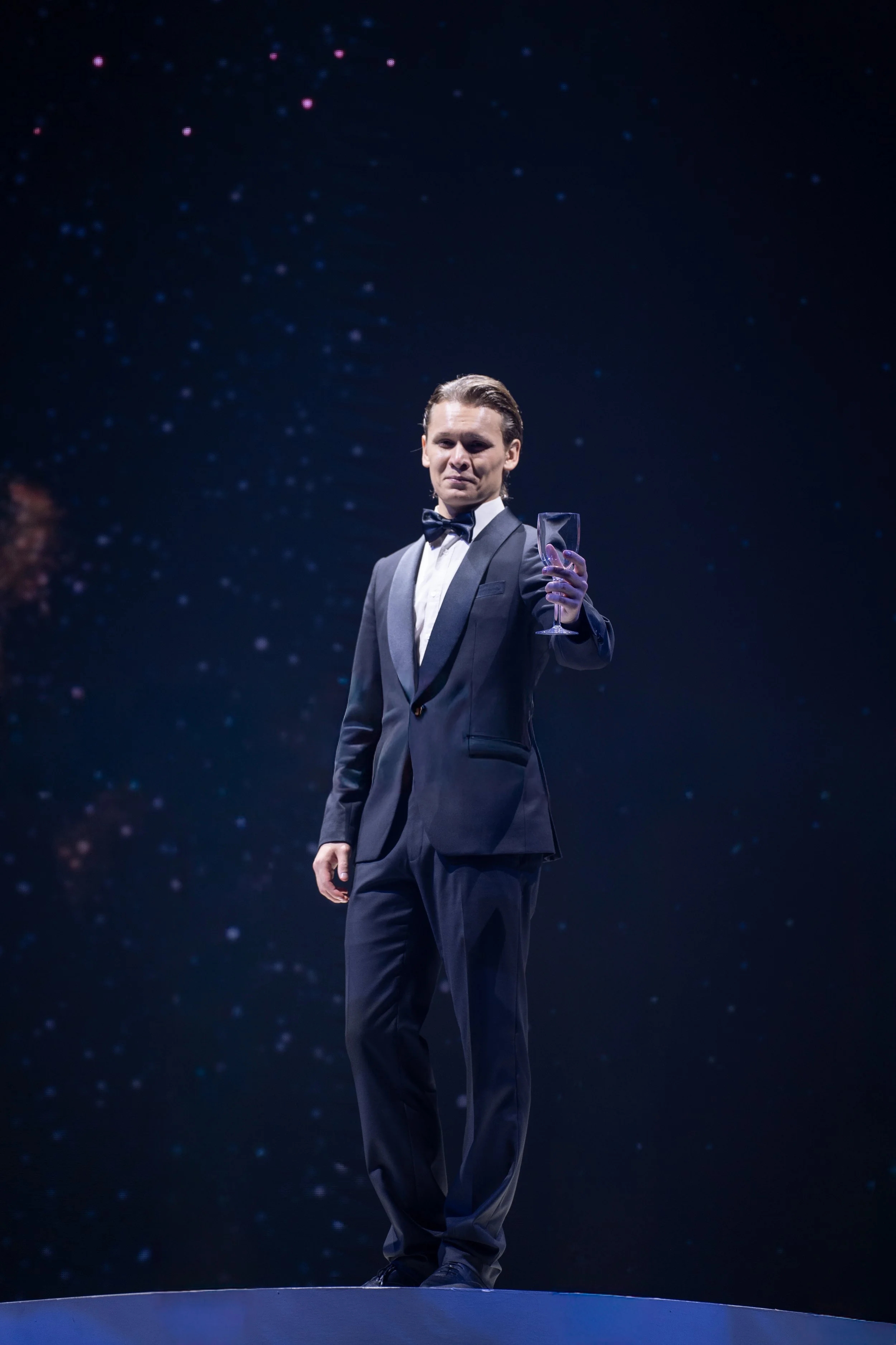 A man in a tuxedo holding a glass of wine in a dark, starry background.