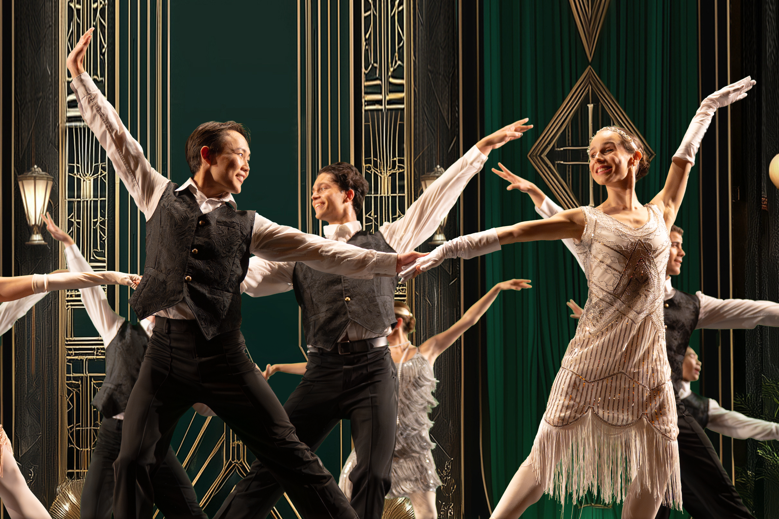 Dancers performing a synchronized routine on stage, dressed in period costumes, with ornate gold and dark green background.