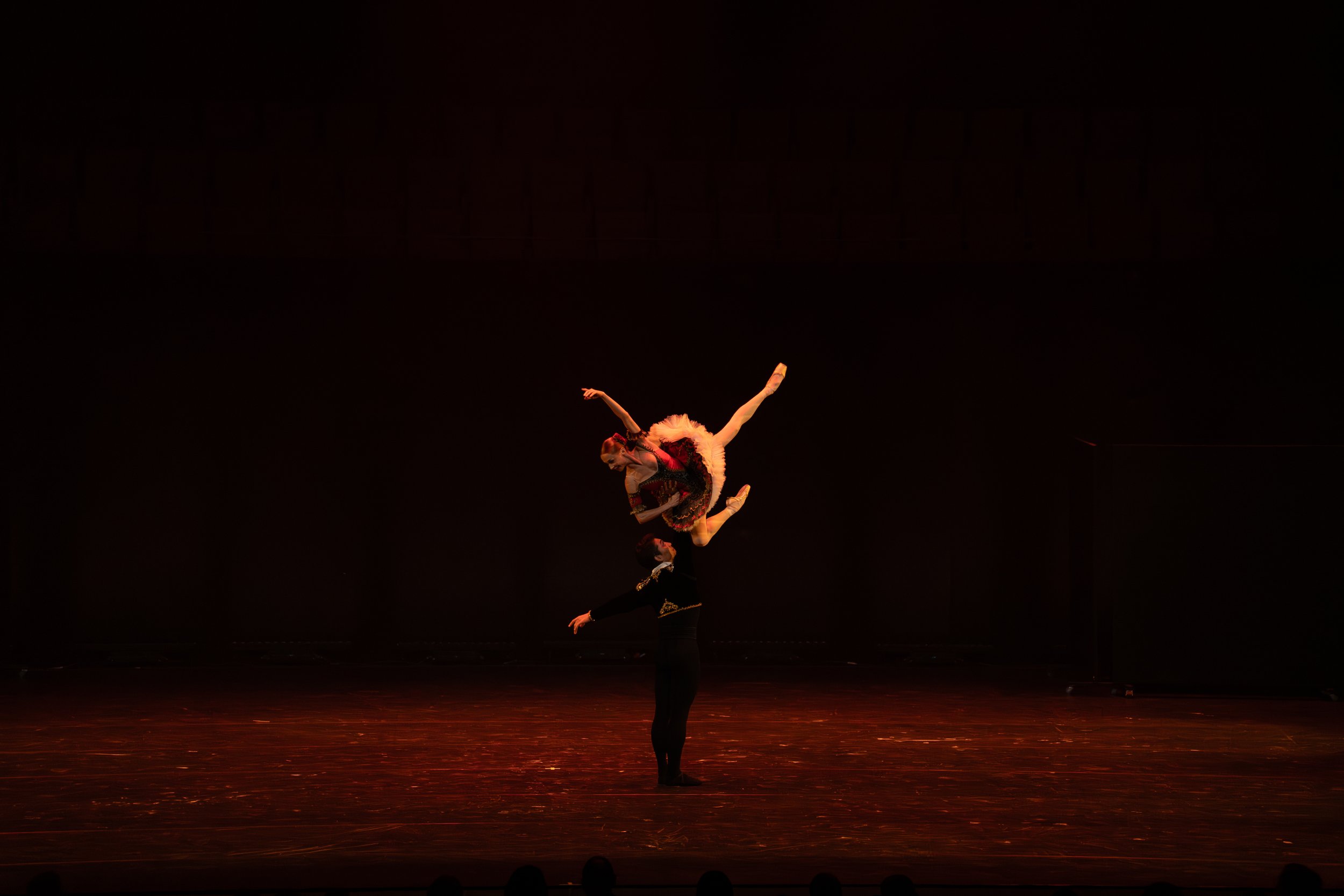 Two performers on stage, one standing with arms raised and another executing an acrobatic pose, illuminated by warm lighting against a dark background.