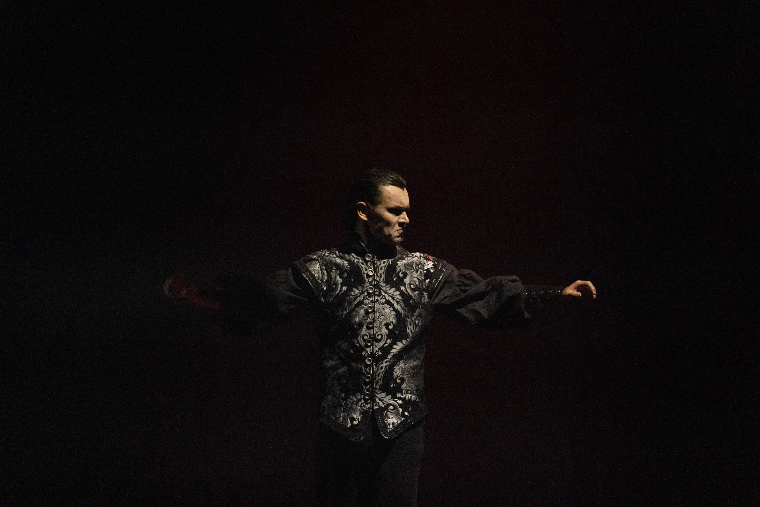 A person in dark ornate clothing with long sleeves stands against a black background, with one arm extended outward.