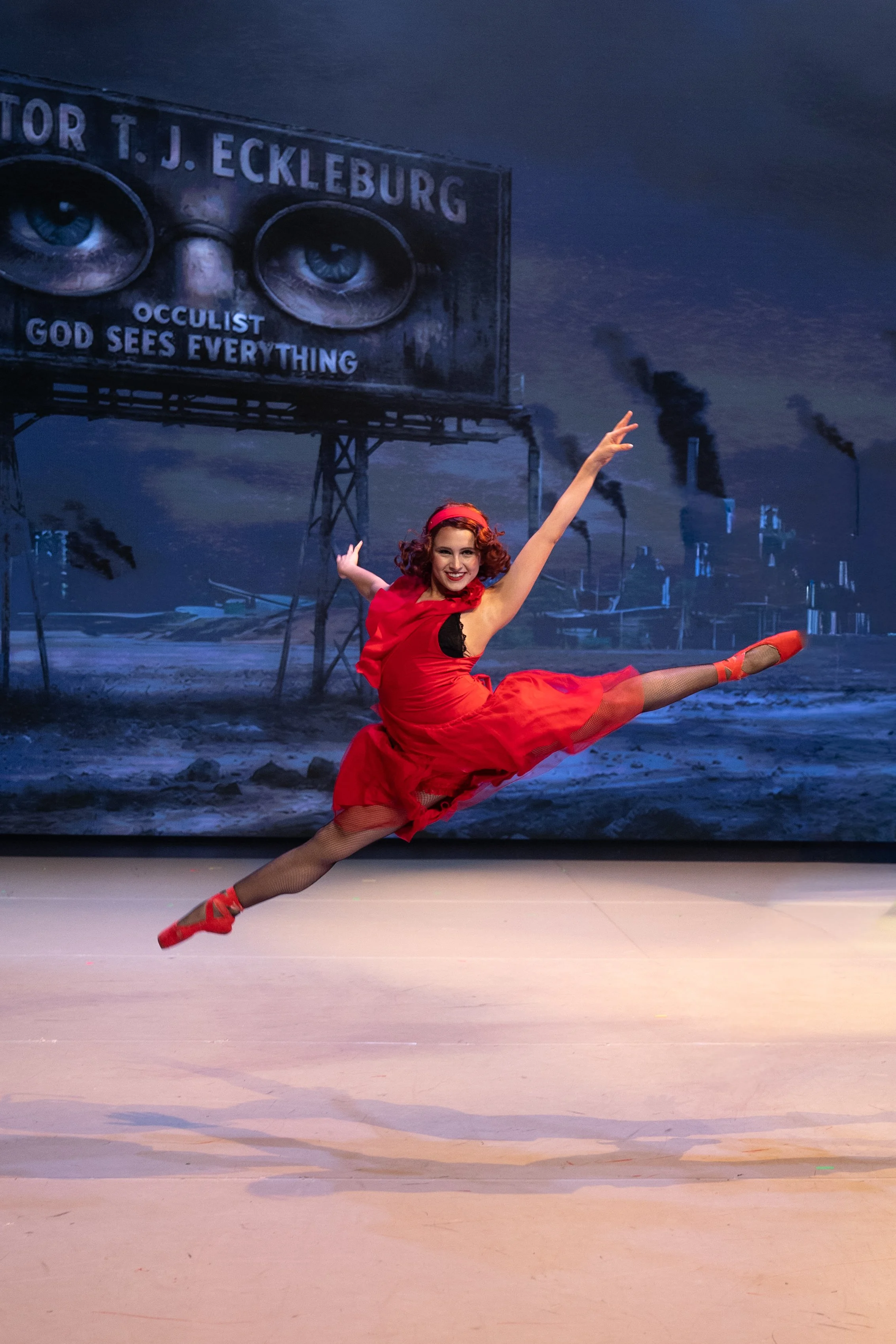 A female ballet dancer in a red dress performing a grand jeté on stage with a dark background depicting an industrial scene and a billboard with eyes and the words 'T J. ECKLEBURG' and 'GOD SEES EVERYTHING'.