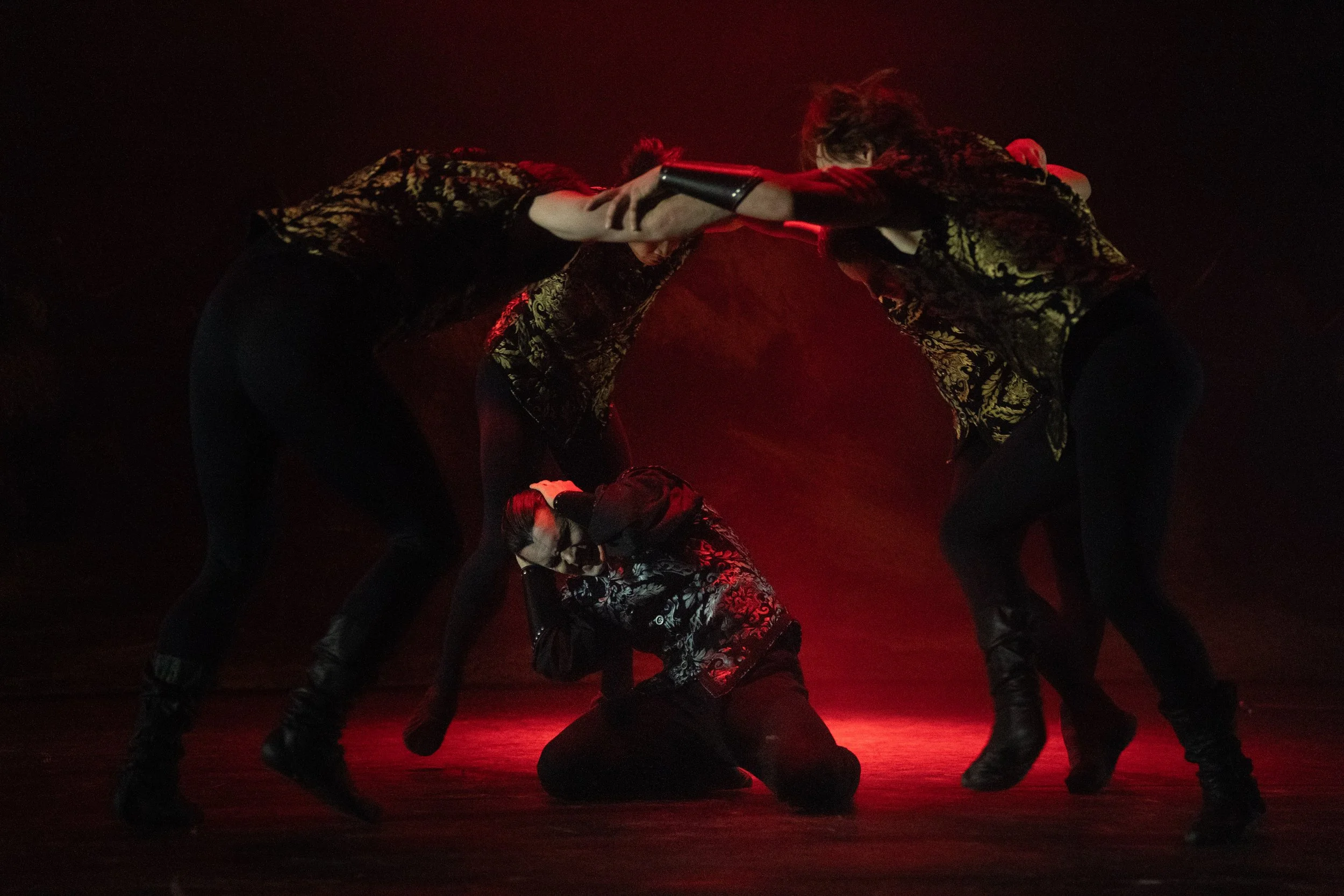 Six performers in dark, ornate costumes with gold patterns, performing a choreographed dance or pose on a dark stage with red lighting in the background.
