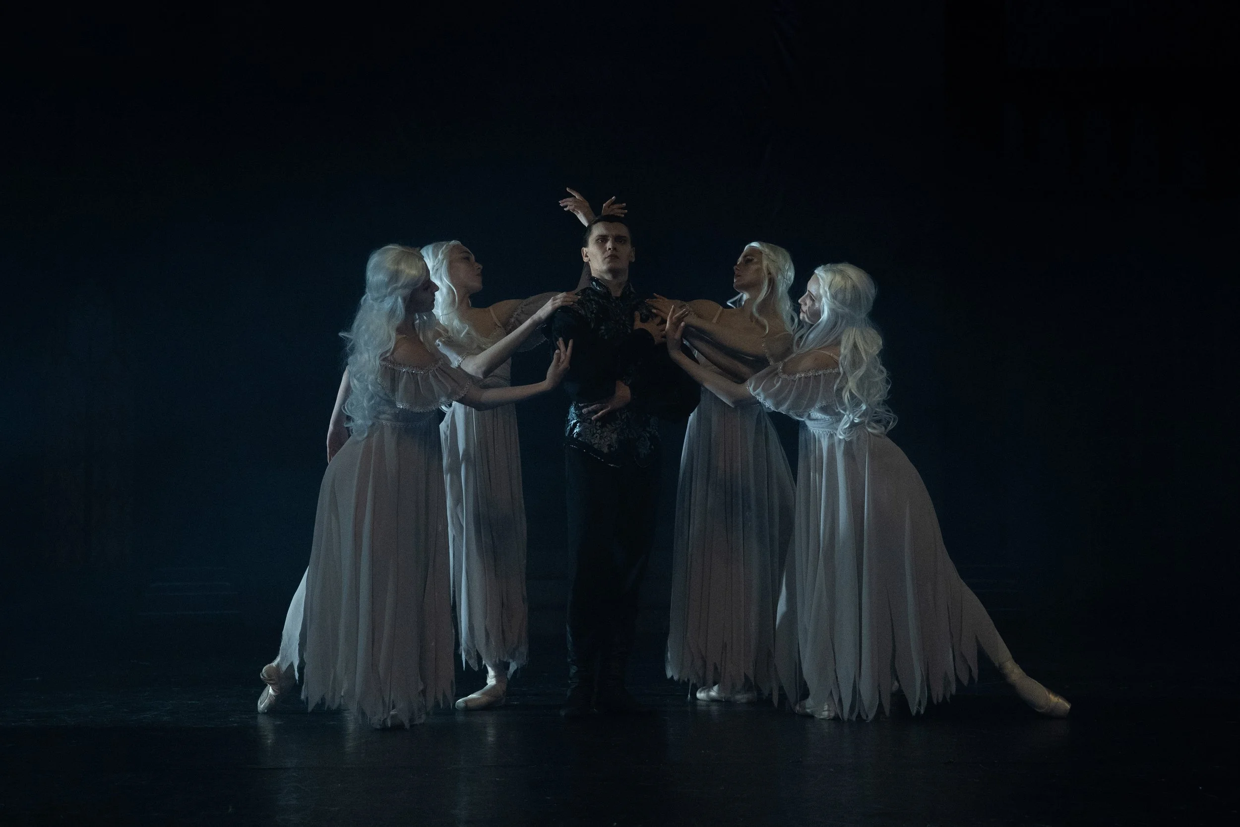 A male ballet dancer in dark clothing surrounded by four female ballet dancers with long white hair, wearing beige costumes, standing in a circle and holding his shoulders and hands, in a dimly lit stage setting.