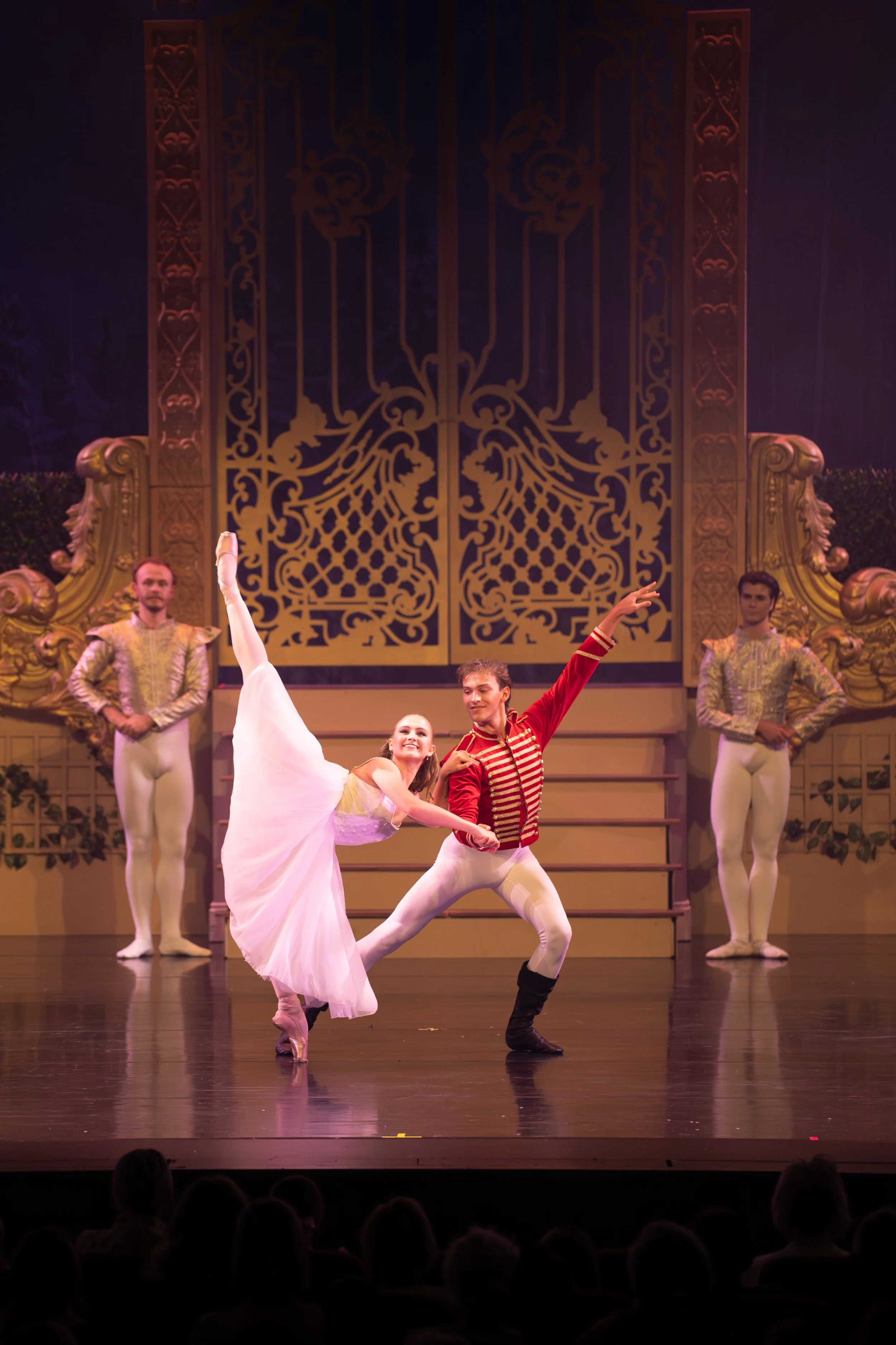 Ballet dancers performing on stage with ornate gold and black backdrop and dressed in costumes resembling 19th-century European style.