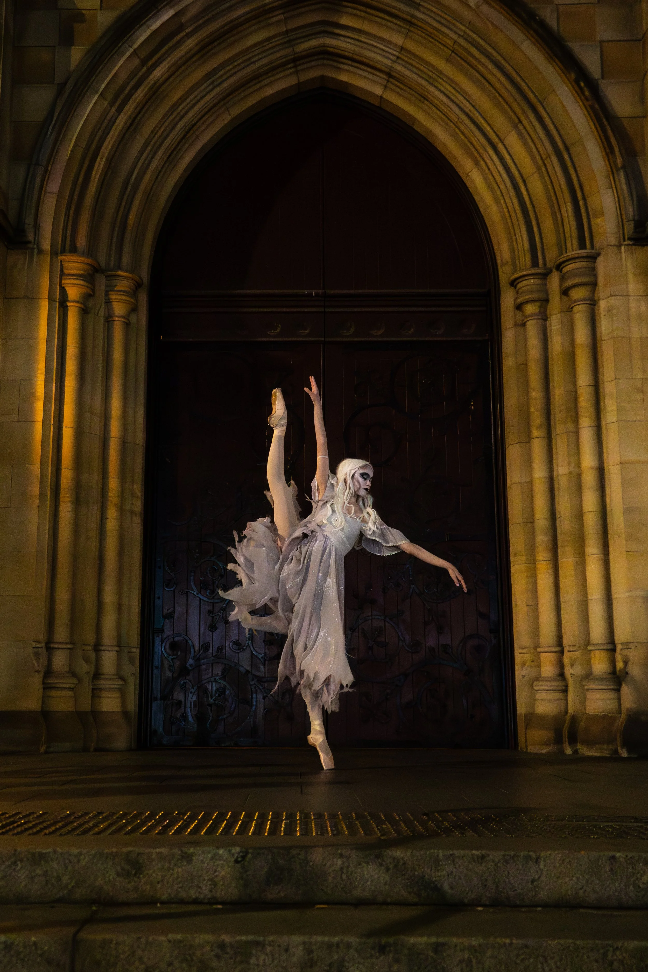 A female ballet dancer is performing a pose in front of a large arched wooden door framed by stone columns, at night. She is dressed in a flowing, light-colored ballet costume.