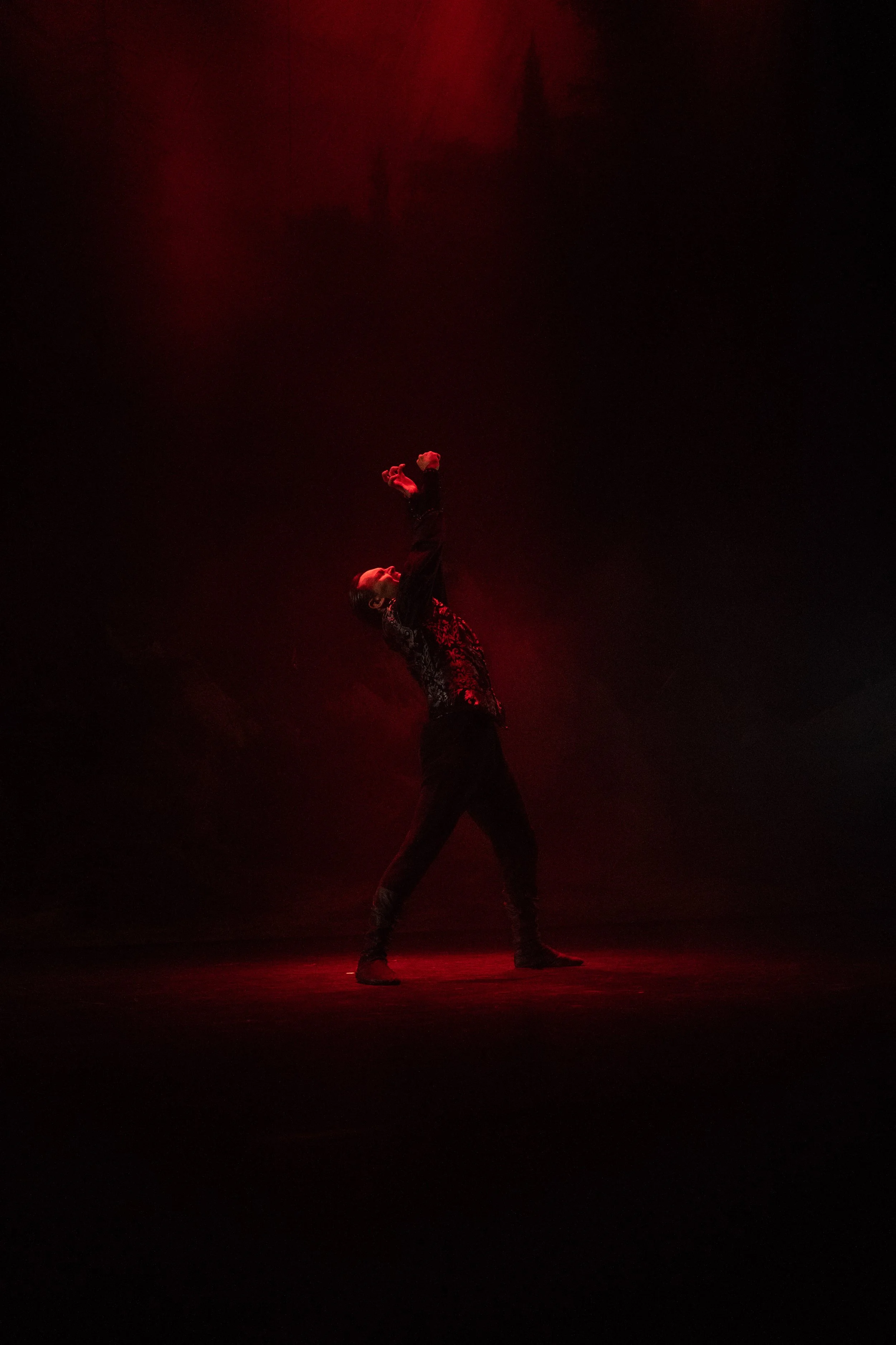 A performer on stage, illuminated by red lighting, in a dynamic dance pose with one arm raised and one arm lowered, wearing dark clothing and boots.