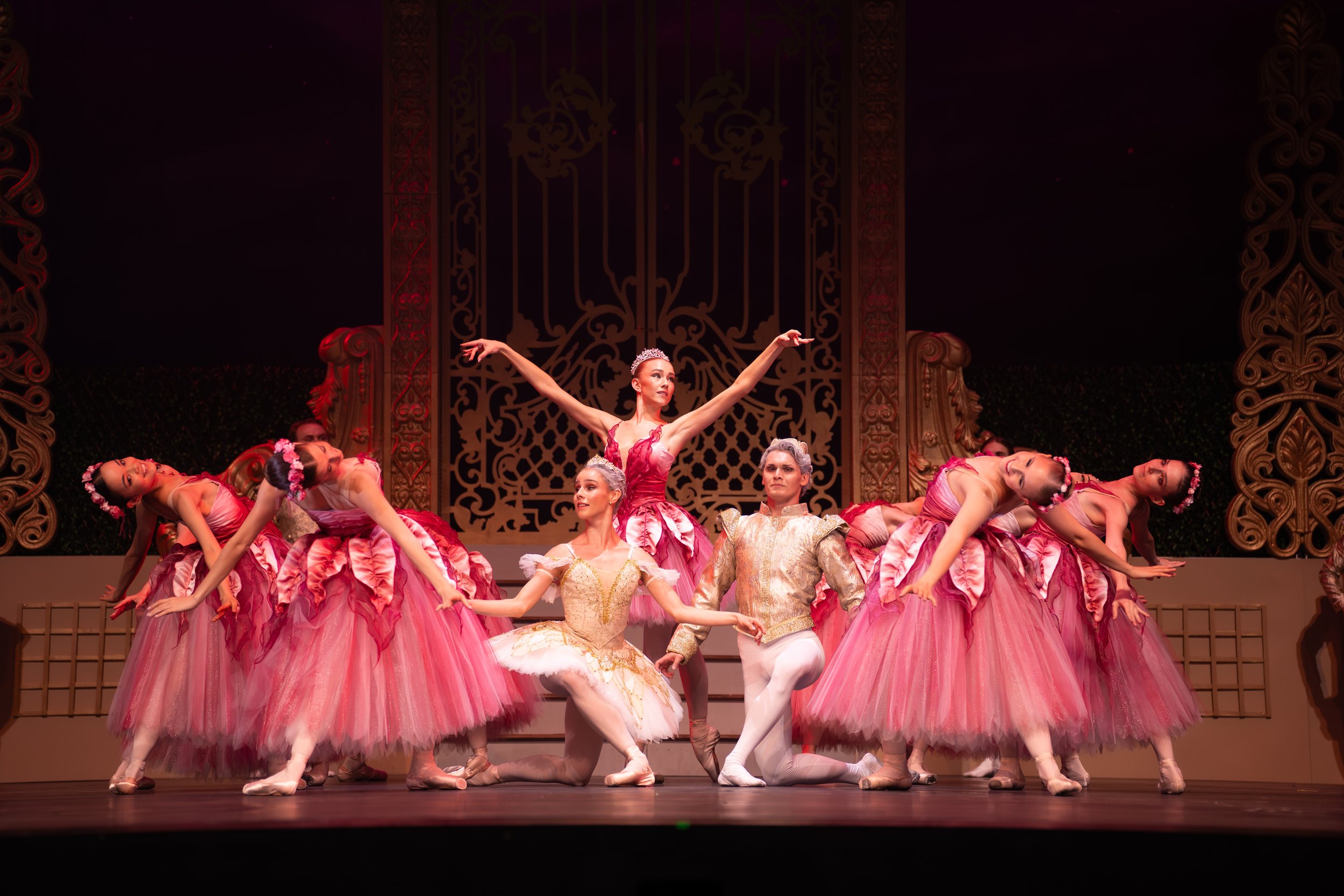 Ballet dancers on stage in elaborate pink costumes with gold accents, performing in a scene with ornate gold and black background decor.