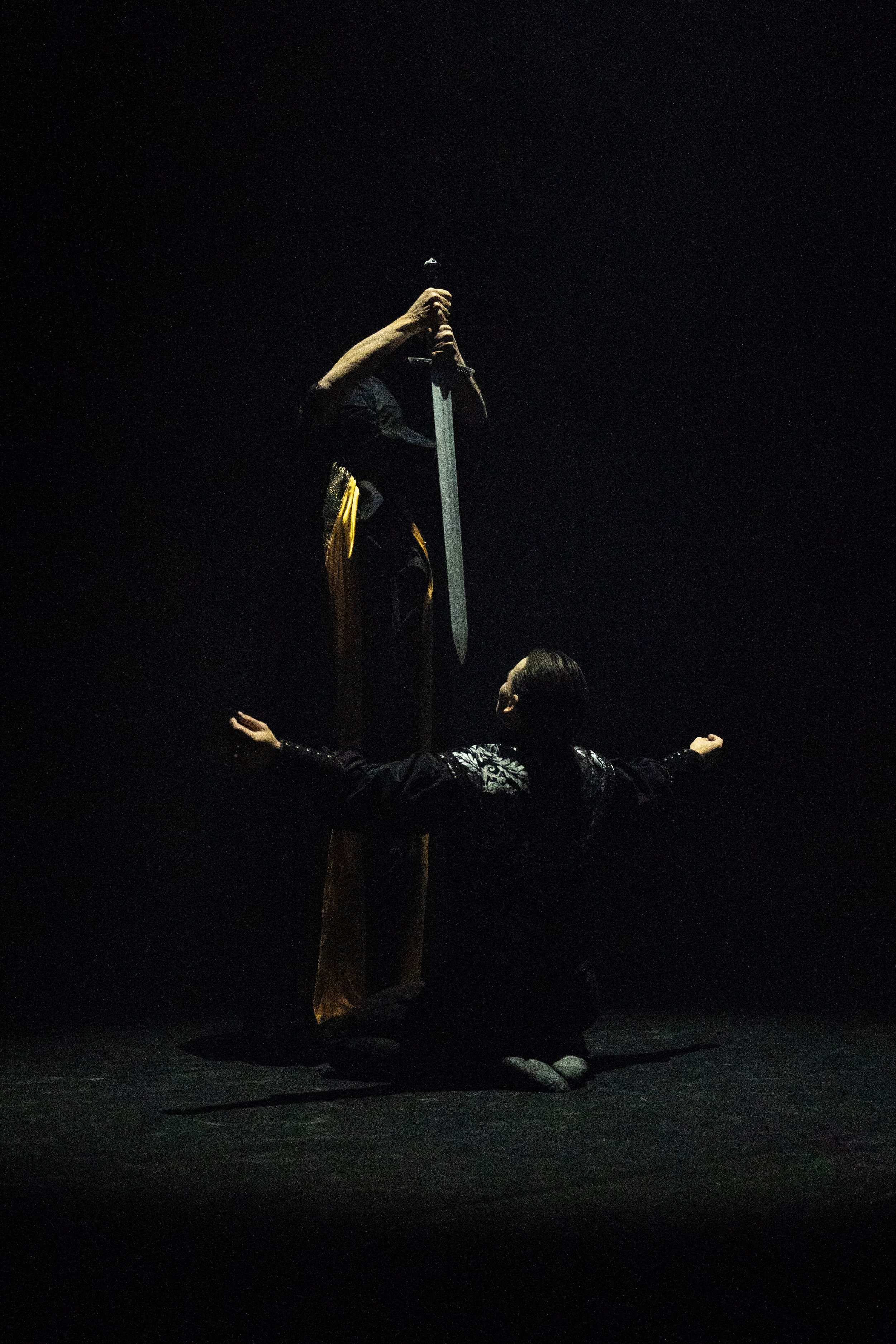 A person kneeling on stage with arms outstretched, another person standing in front with hands on shoulders, and a third person in the background. The person standing is holding a sword vertically, with a dark background.