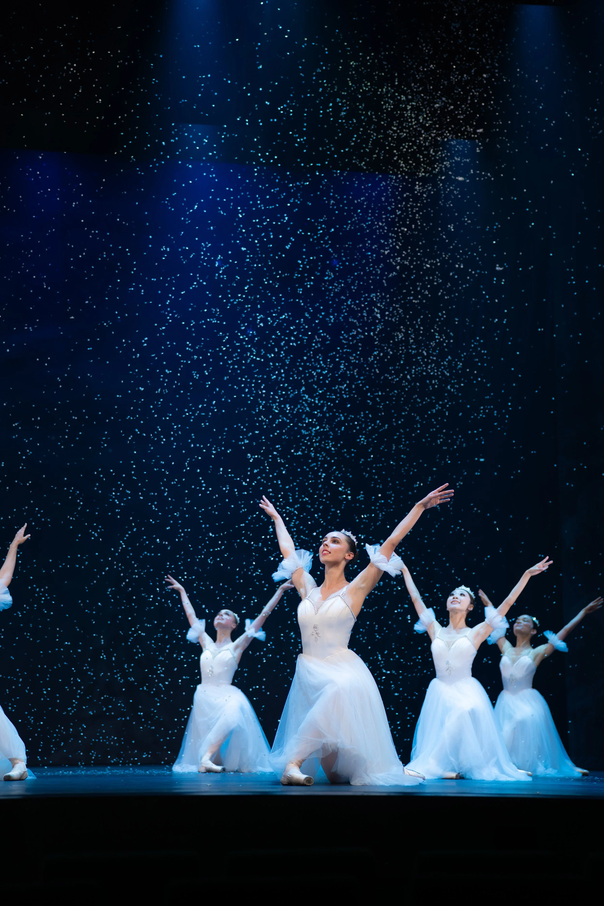 Ballet dancers in white costumes performing on stage with dark background and falling confetti.