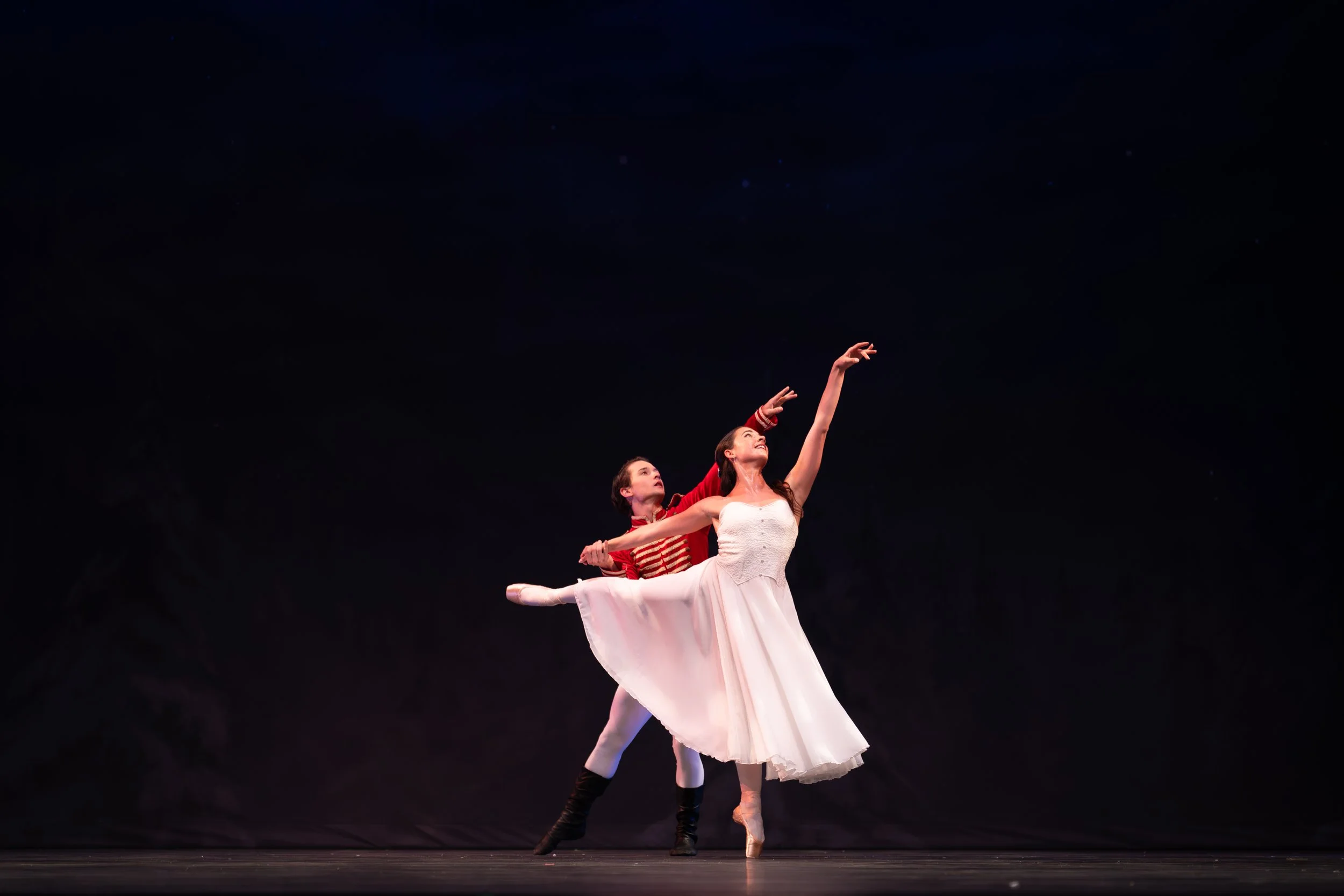 Two ballet dancers perform on stage against a dark backdrop. One dancer stands en pointe with her right arm extended upward, wearing a white tutu. The other dancer, wearing a red and white striped shirt and black leggings, supports her from behind wi