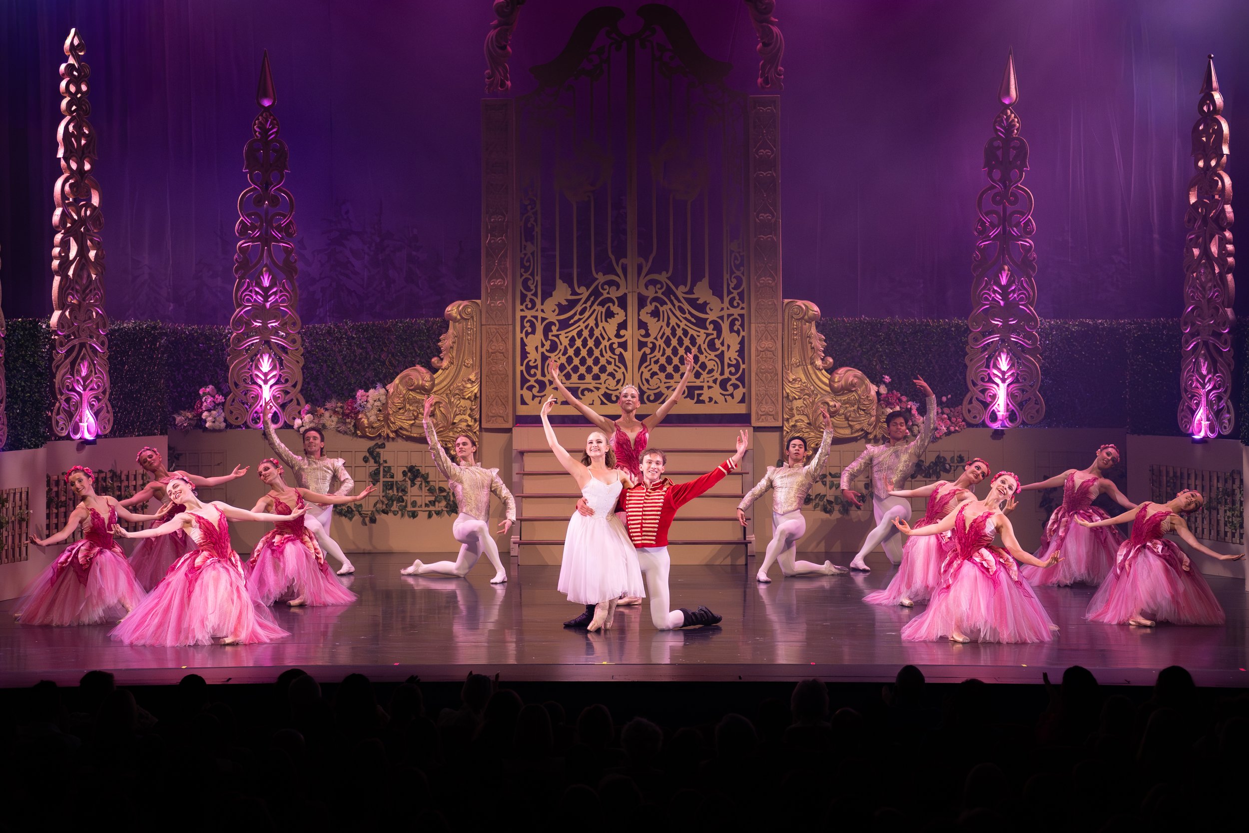 Ballet dancers performing on stage in elaborate costumes with pink tutus, a male and female lead in the center, surrounded by other dancers, set against a backdrop with ornate gates and purple lighting.