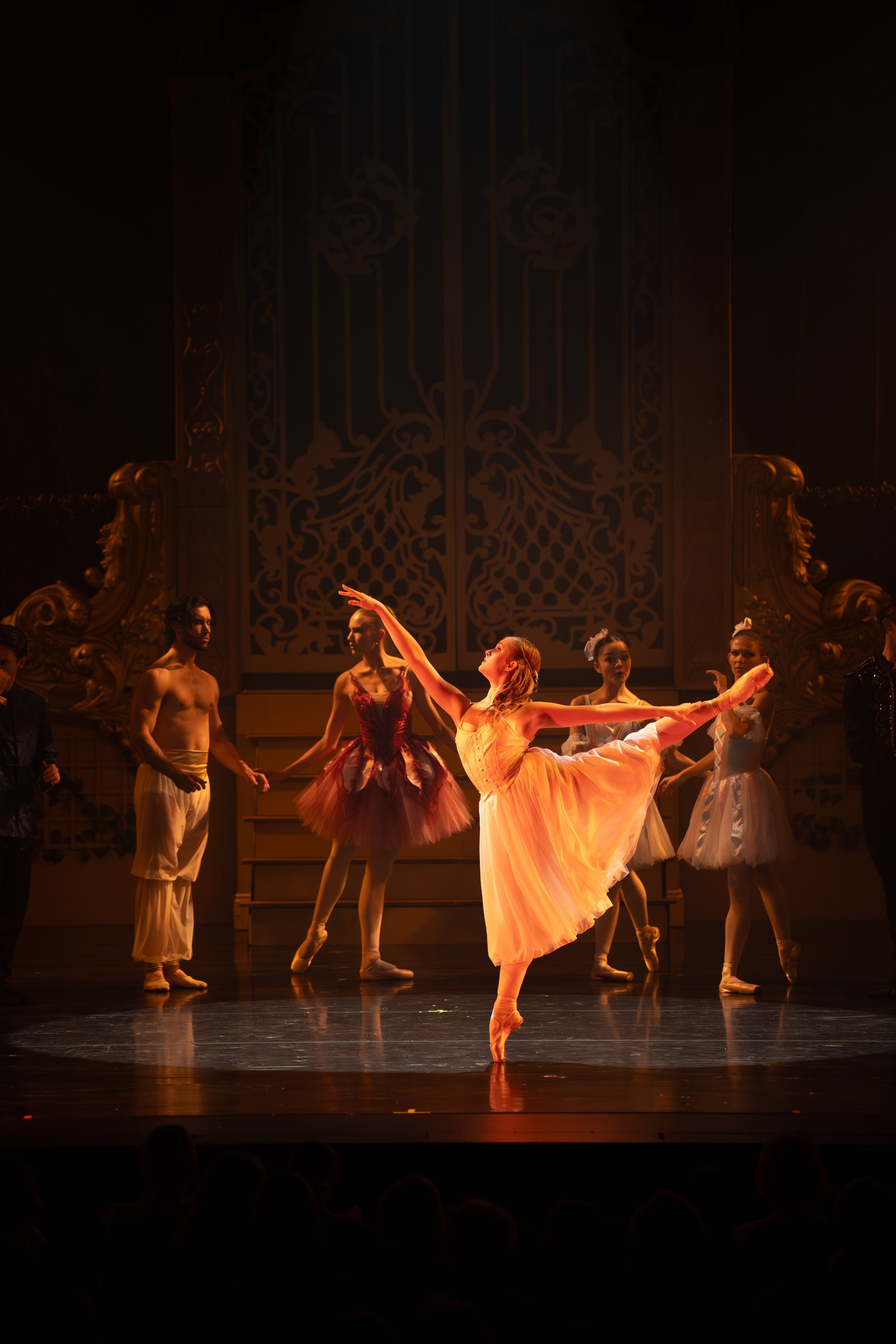 Ballet dancers performing on stage with a grand ornate backdrop, one dancer in a yellow dress is in a graceful pose at the center.