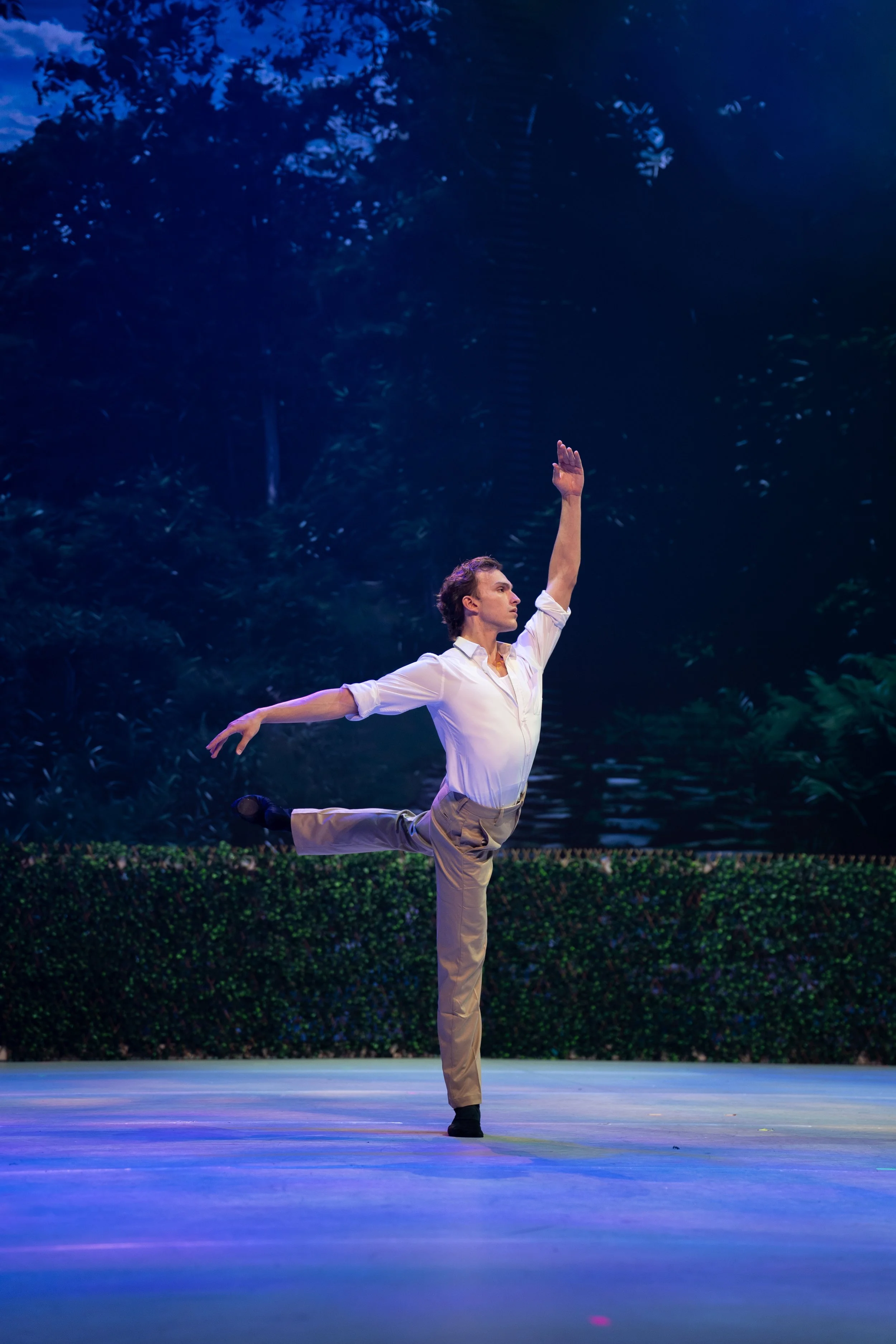 A ballet dancer in a white shirt and beige pants performs a pose on stage, with one arm raised and the other arm extended to the side, in front of a background resembling a lush forest scene.