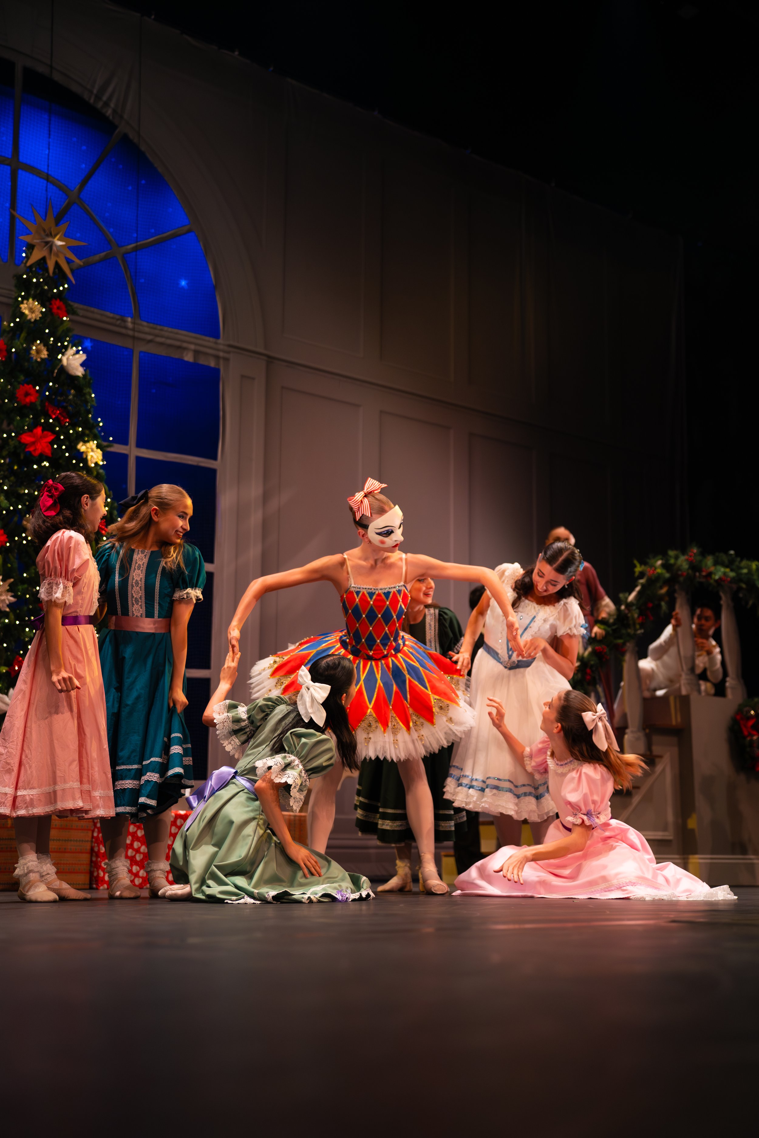 Scene from a ballet performance with dancers in colorful costumes on stage, Christmas tree decorated with ornaments and lights, large window in the background, and ballerinas in various poses, one wearing a harlequin costume.