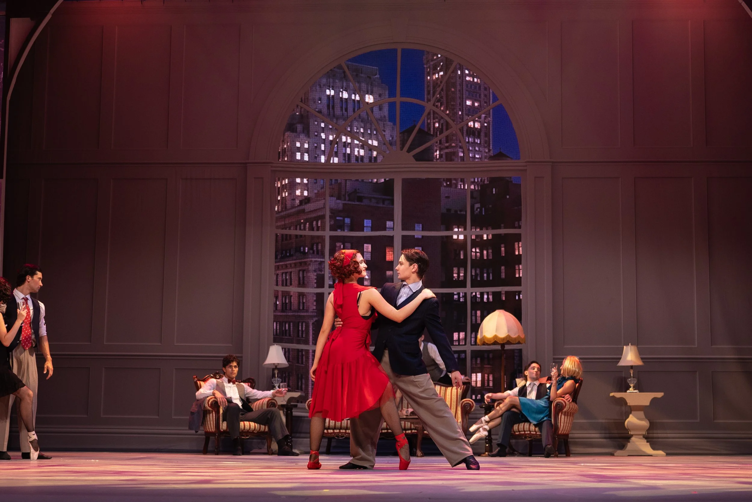 Actors performing a dance on stage with a cityscape backdrop at night, some sitting and others standing or dancing, including a woman in a red dress and a man in a suit.