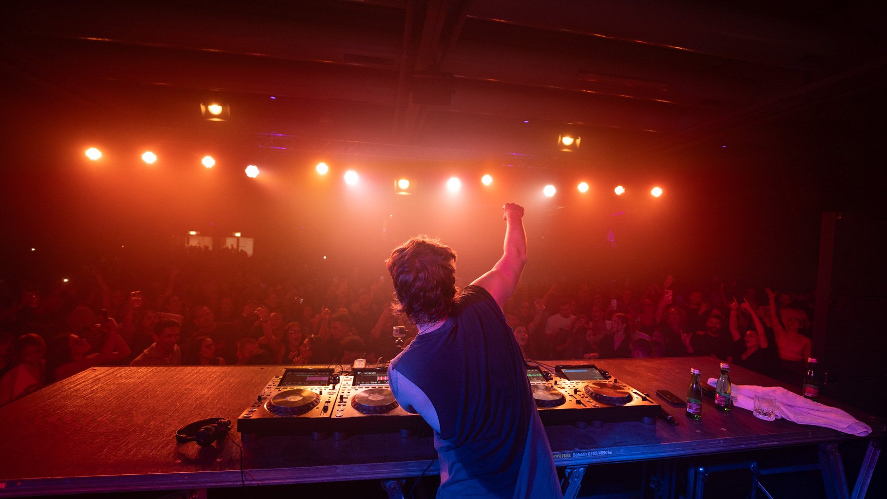 DJ performt auf einer Bühne in einem Club, die Menge feiert und hebt die Arme, im Hintergrund warme Bühnenlichter.