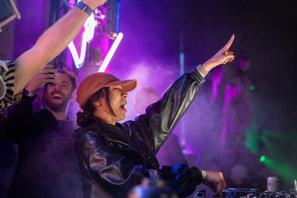 Junge Frau als DJ im Club, die mit den Armen in die Luft streckt, umgeben von tanzenden Leuten bei bunten Lichteffekten.