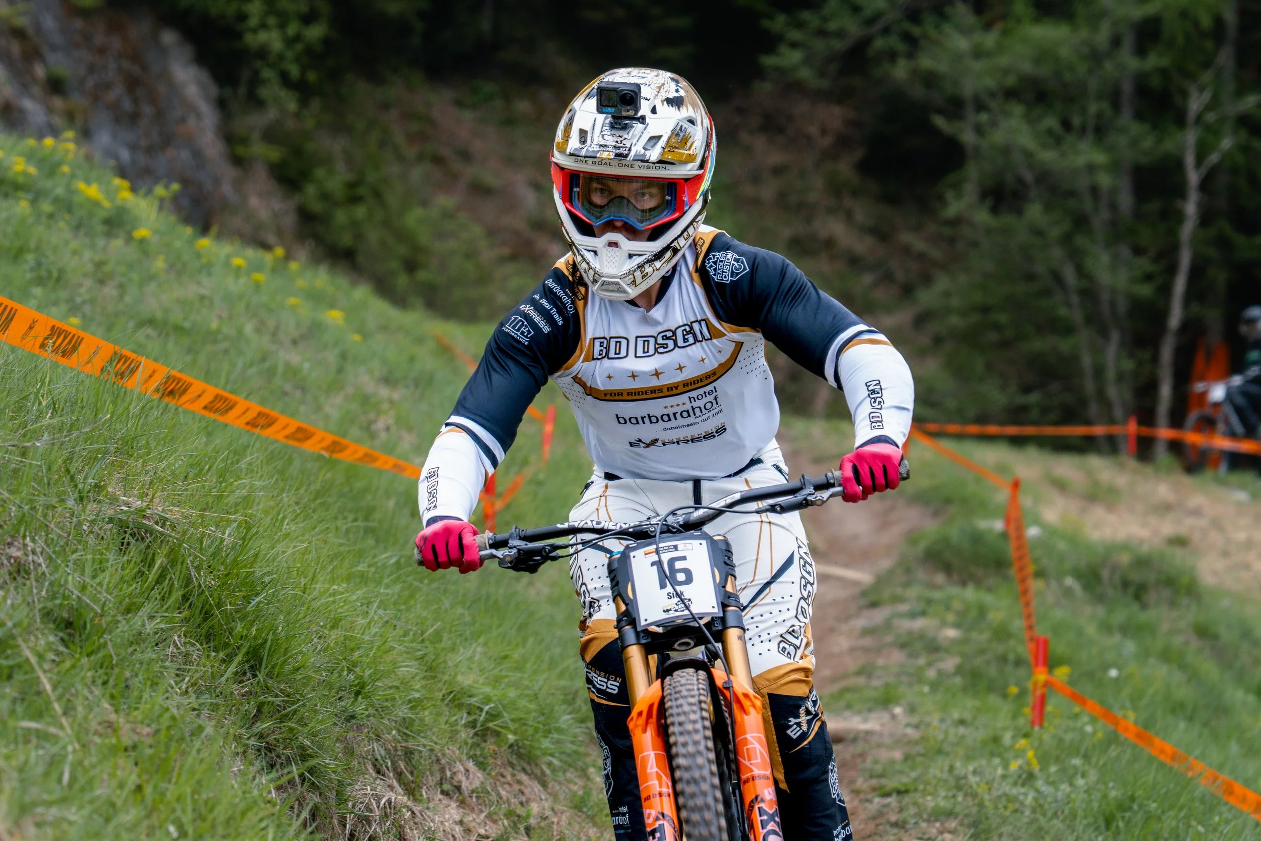 Mountainbiker in Schutzhelm und Radhandschuhen beim Rennen auf einem waldigen Trail, umgeben von grünem Gras und Bäumen, mit orangefarbener Absperrung.