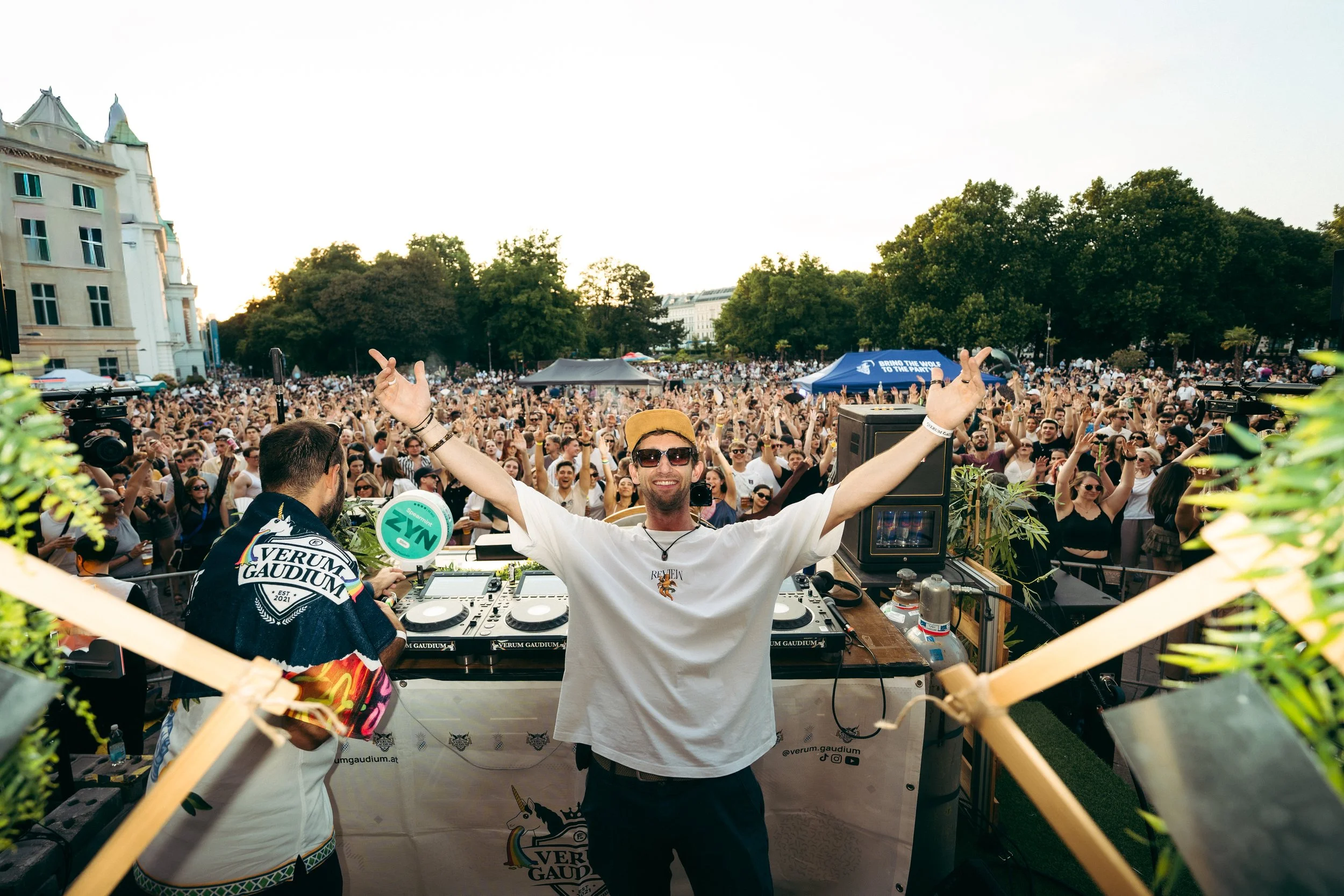 DJ steht hinter einem Mischpult auf einer großen Menschenmenge bei einem Open-Air-Festival, glücklicher Mann mit Sonnenbrille und gelber Mütze im Vordergrund, viele Fans jubeln und tanzen im Hintergrund, zahlreiche Bäume und Gebäude im Park, tagsüber bei Sonnenschein.