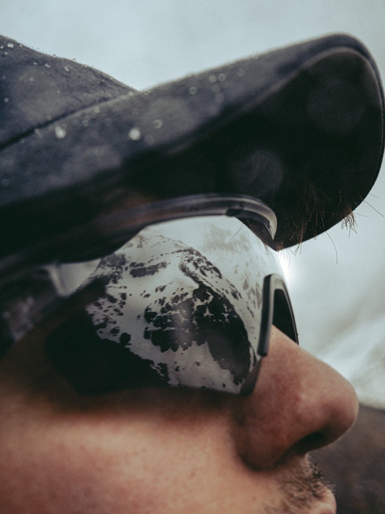 Nahaufnahme einer Person mit Ski- oder Snowboardbrille und Cap, im Hintergrund schneebedeckte Berge.