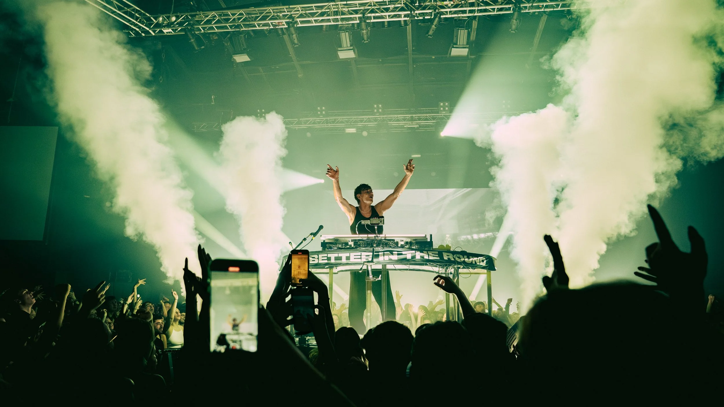DJ performs auf einer Bühne bei einem Konzert mit grüner Beleuchtung und Rauch, vor Publikum mit erhobenen Händen.