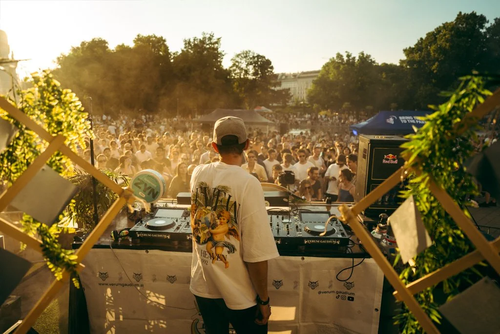 DJ bei einem Freiluft-Festival, blickt auf eine große Menschenmenge, von der Sonne beleuchtet, im Hintergrund Bäume und ein historisches Gebäude.